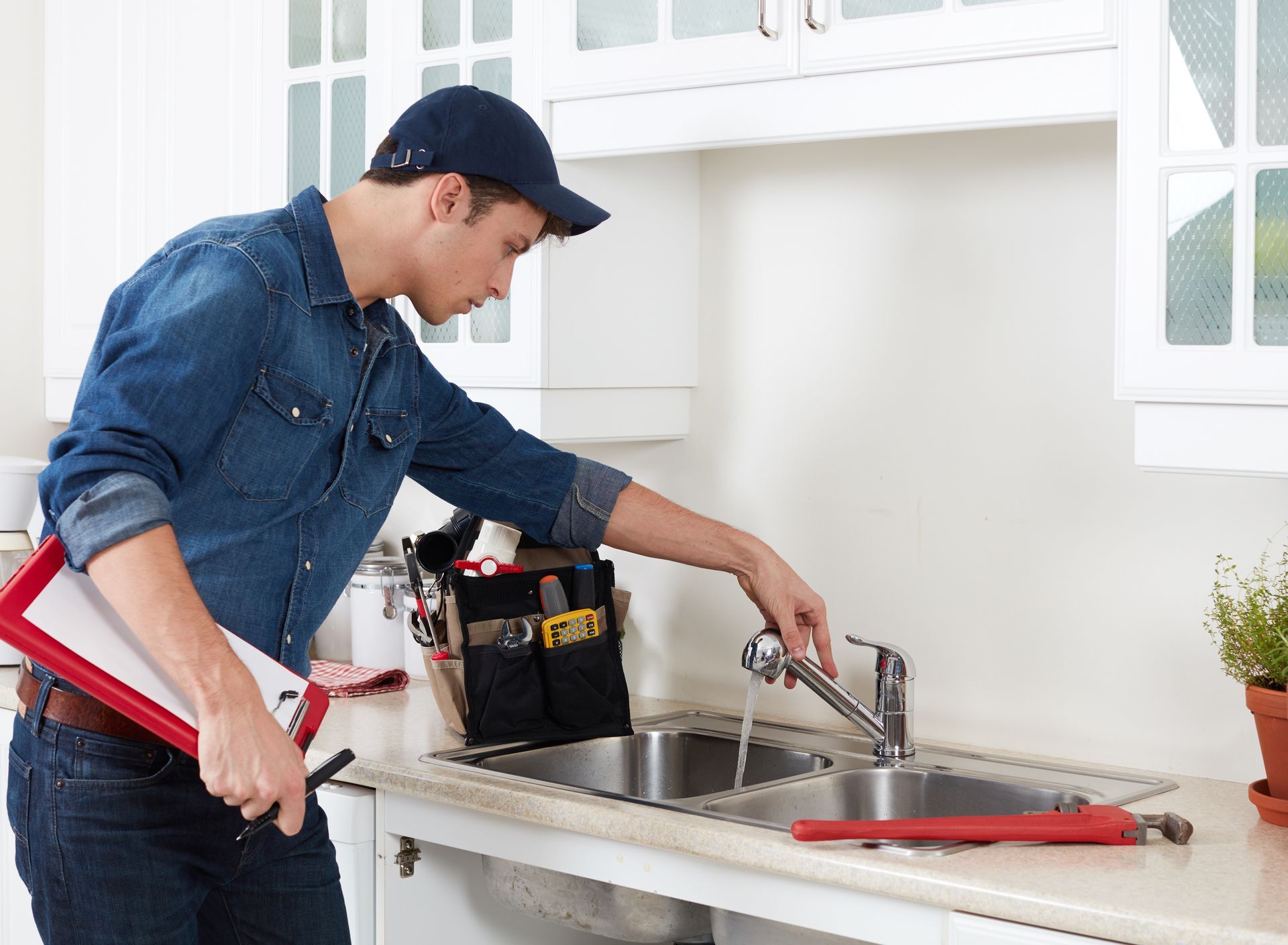 Plumber in denim shirt and hat, examining a kitchen faucet with running water. Tool box and clipboard present.