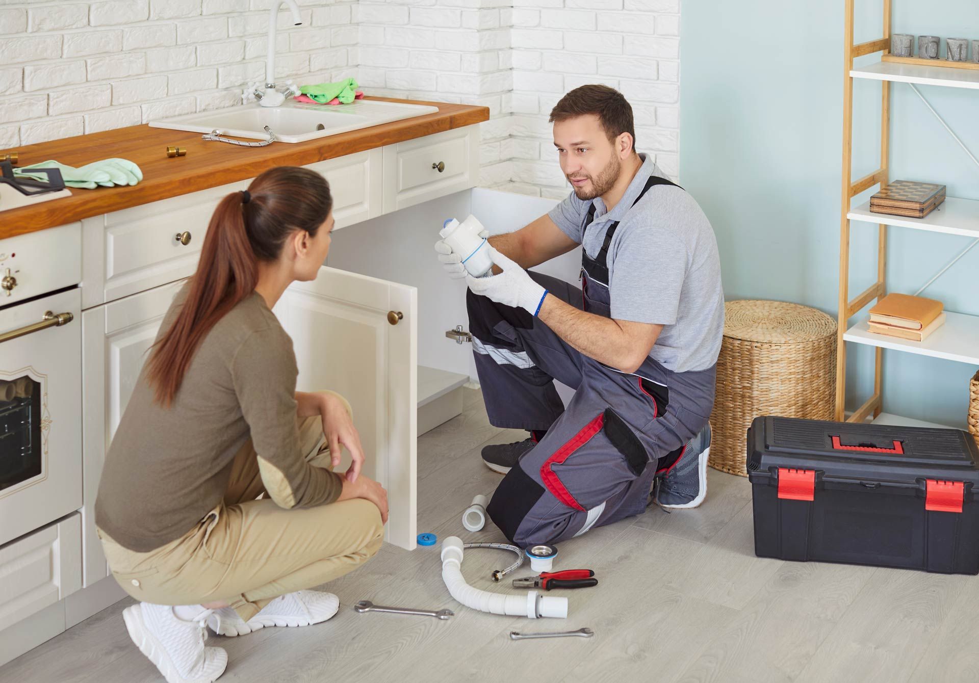 The plumber shows the pipe part while the client observes the repair in the kitchen.