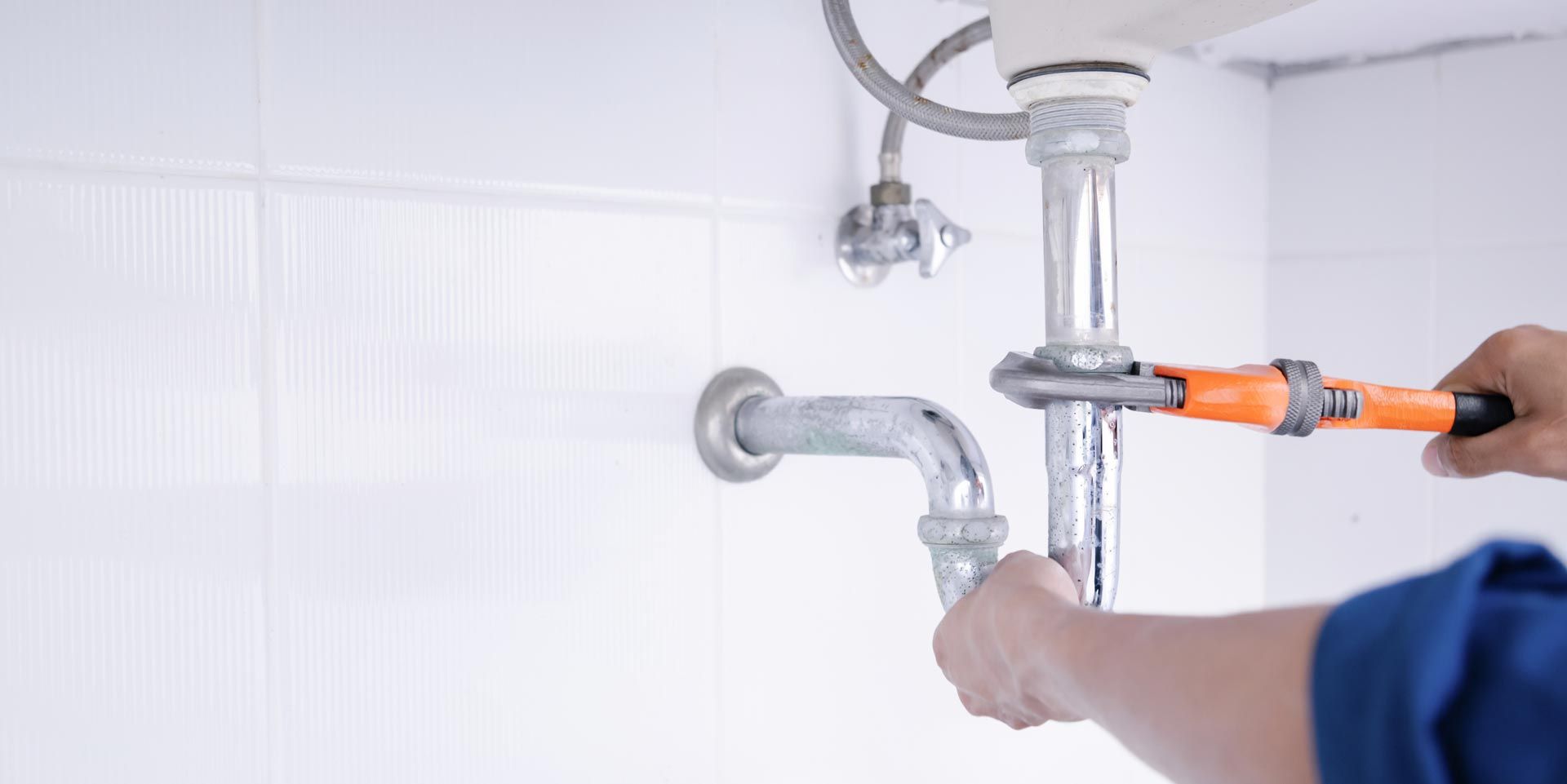 A person using an orange pipe wrench to tighten or loosen a metal pipe under a sink.