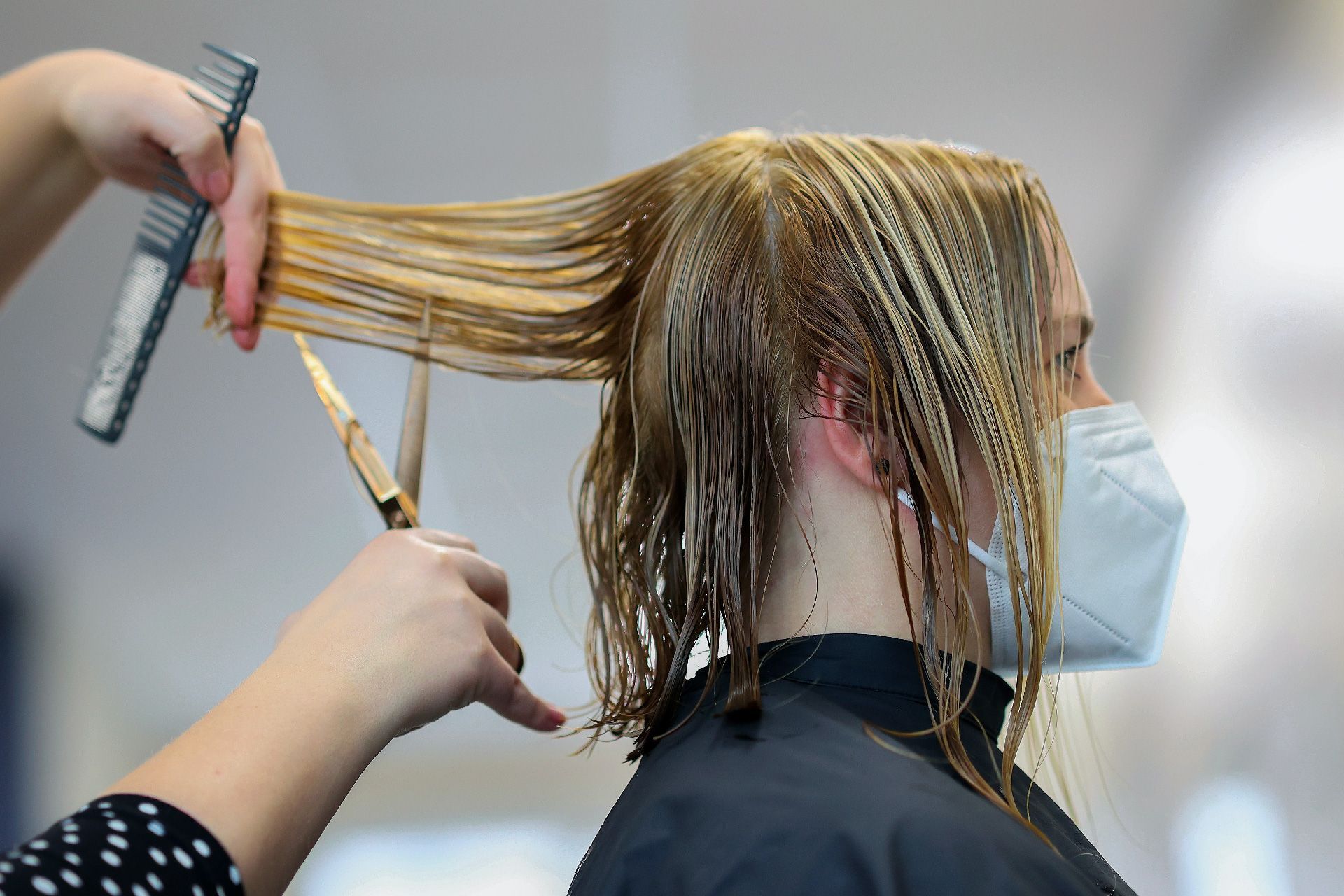 Peluquera cortando el cabello rubio y mojado de una persona con mascarilla. Manos con herramientas, en el interior.