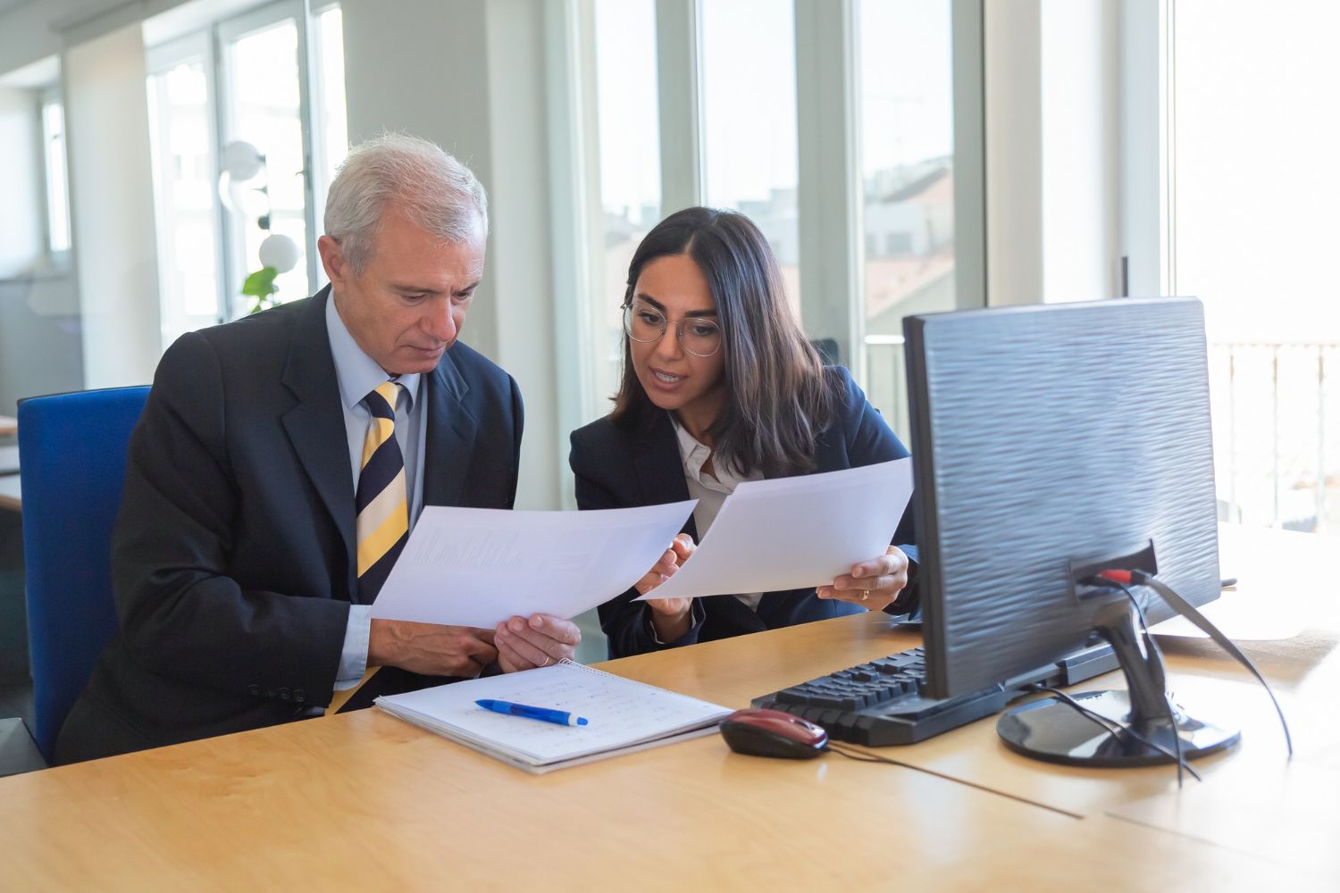 Two professionals in office, looking at papers; man in suit, woman in glasses, desk, computer.