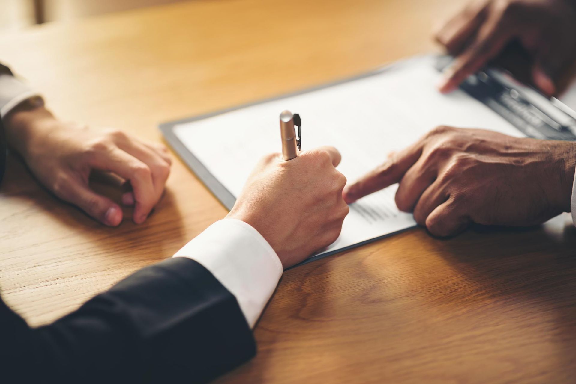 Person signing document, another person pointing at a section on a wooden table.