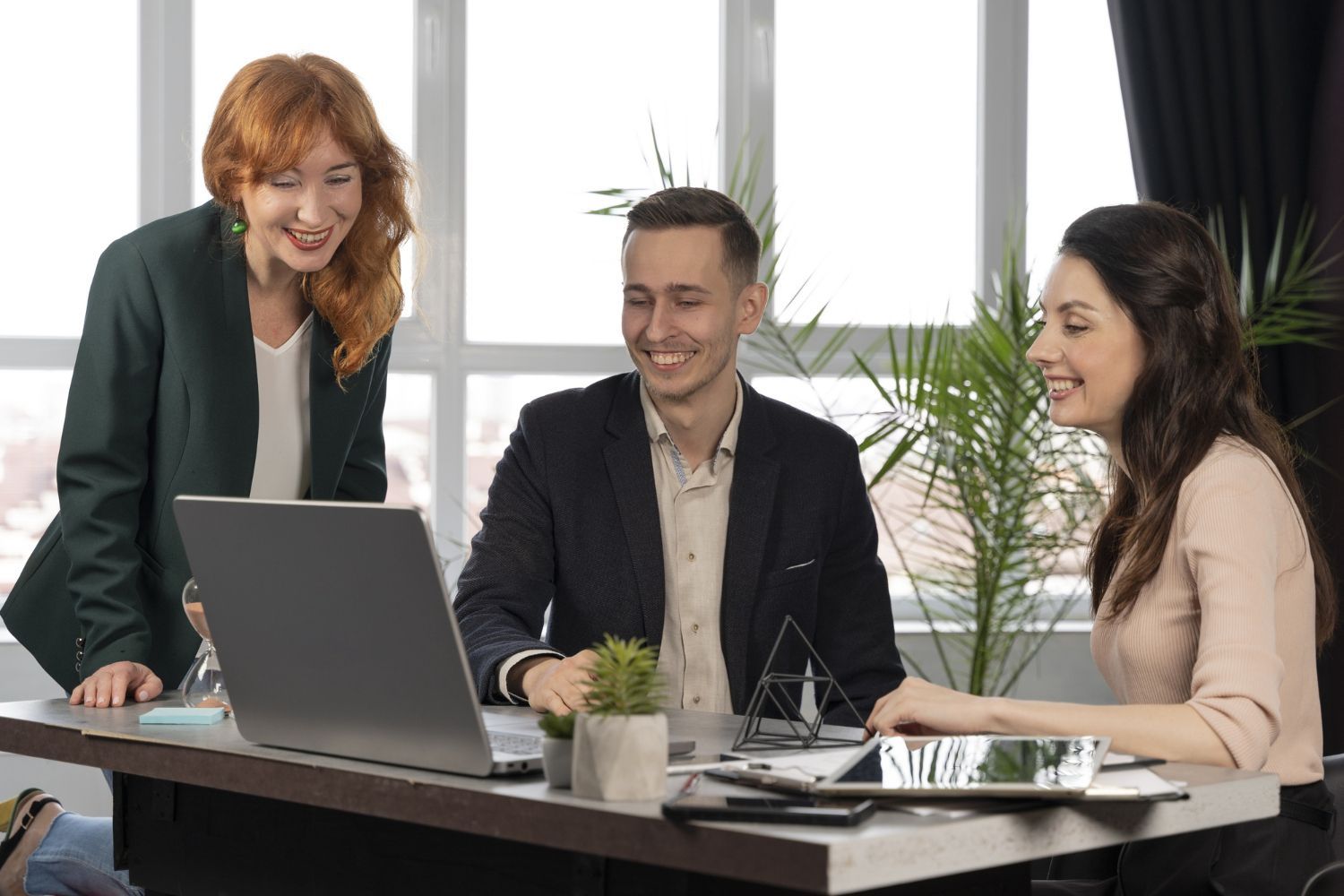 Three people in business attire around a laptop, smiling. Office setting with natural light, a plant, and a tablet.