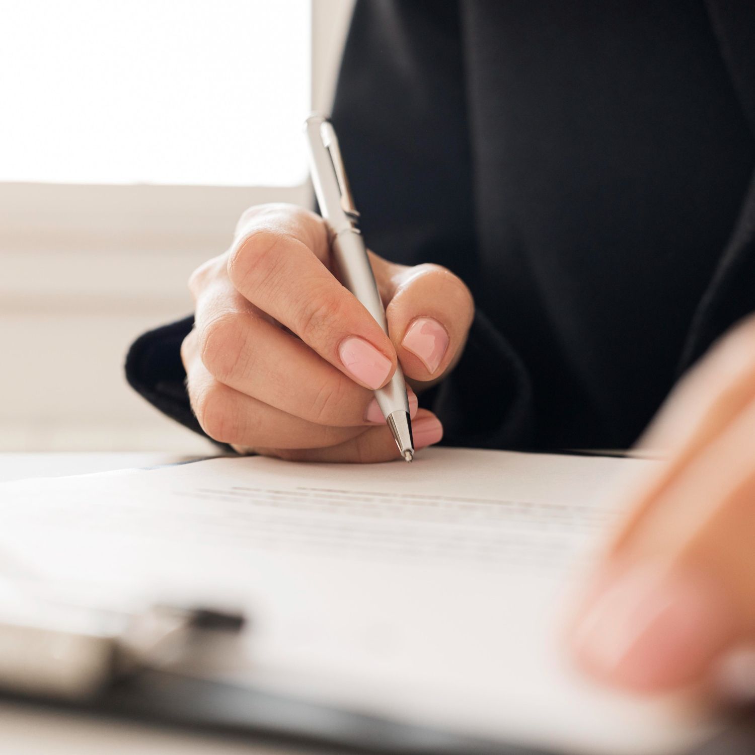 Person writing on a paper with a silver pen, wearing a black blazer.