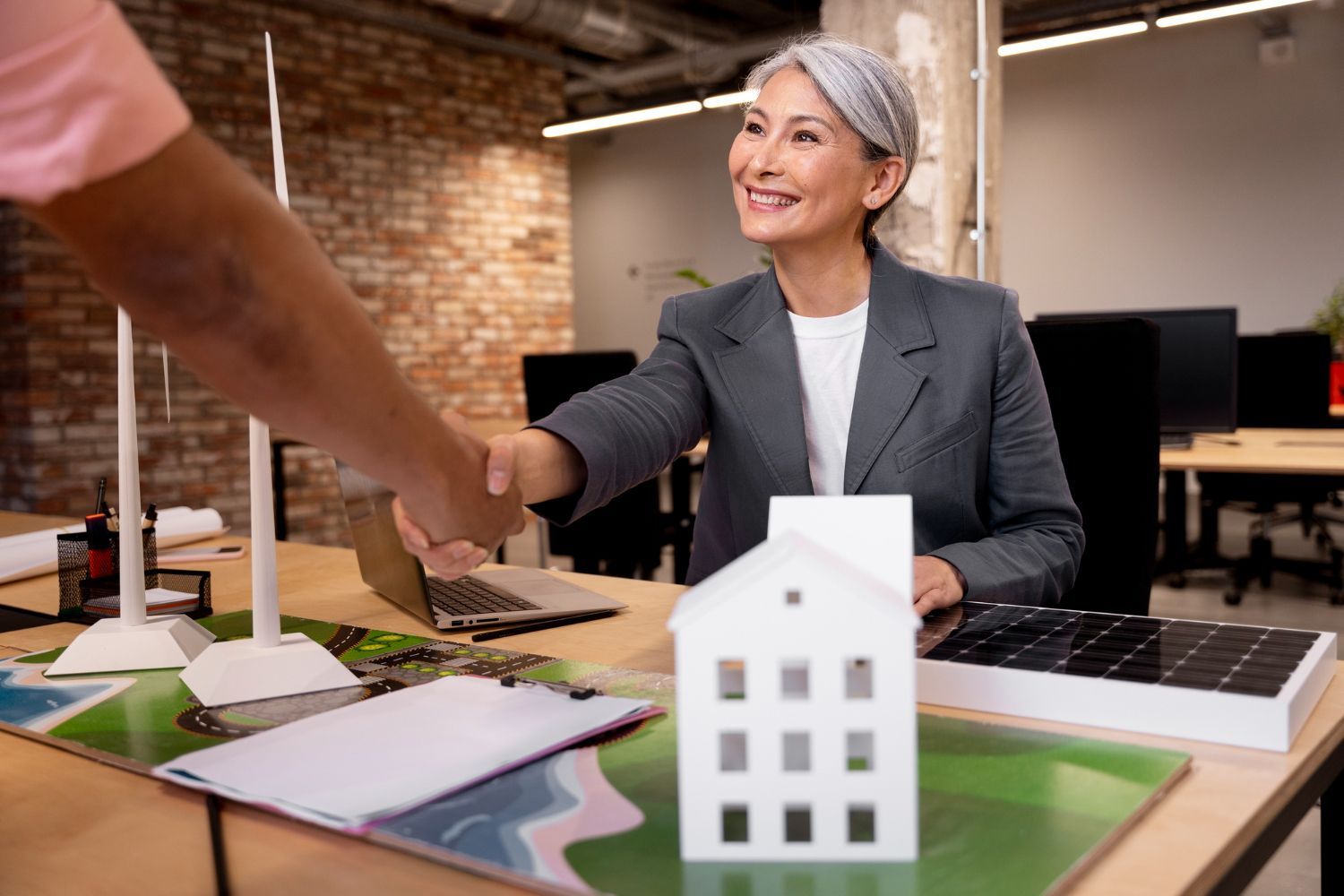 Woman in suit shaking hands at office desk with house model and solar panel.