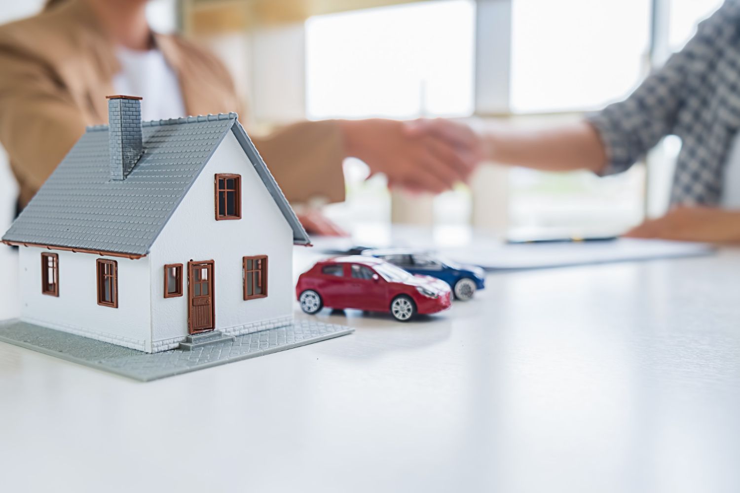 Miniature house and cars on a table with people shaking hands in the background.