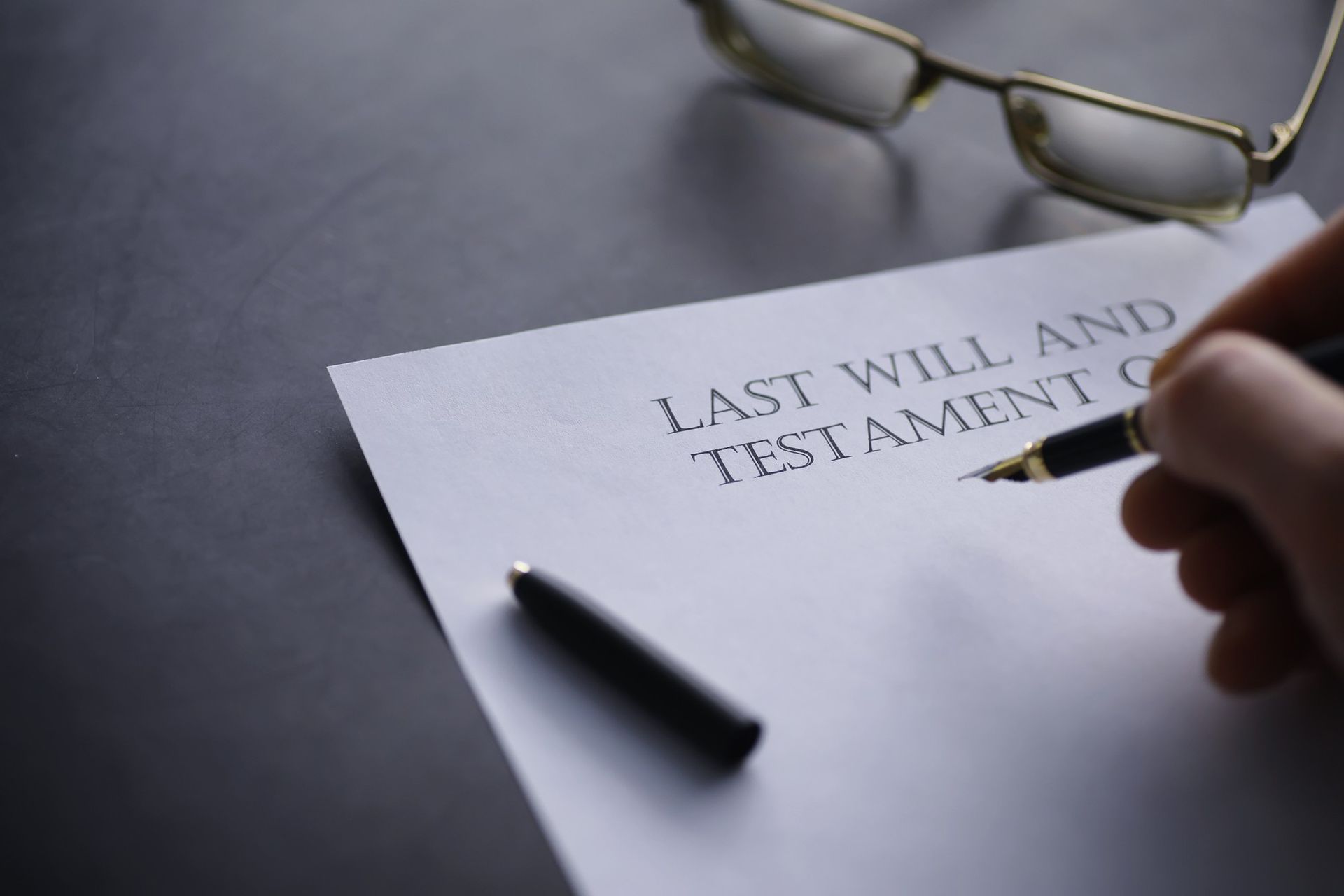A hand signing a Last Will and Testament document, glasses on a dark surface.