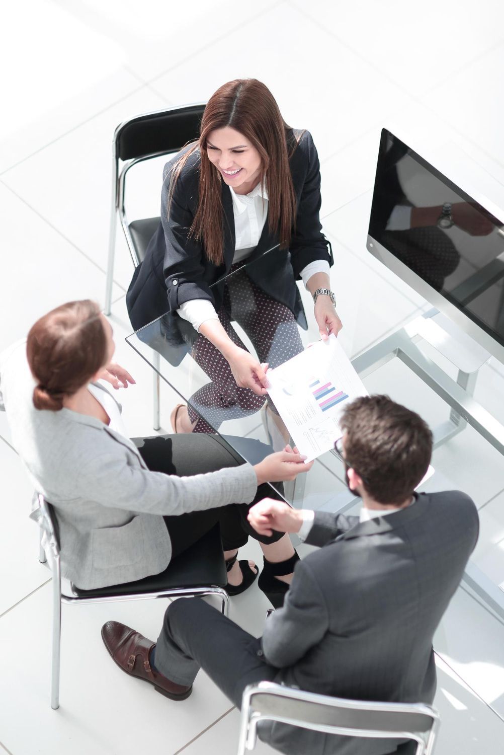Business colleagues reviewing paperwork at a glass table; one woman smiles, handing the paper to others.