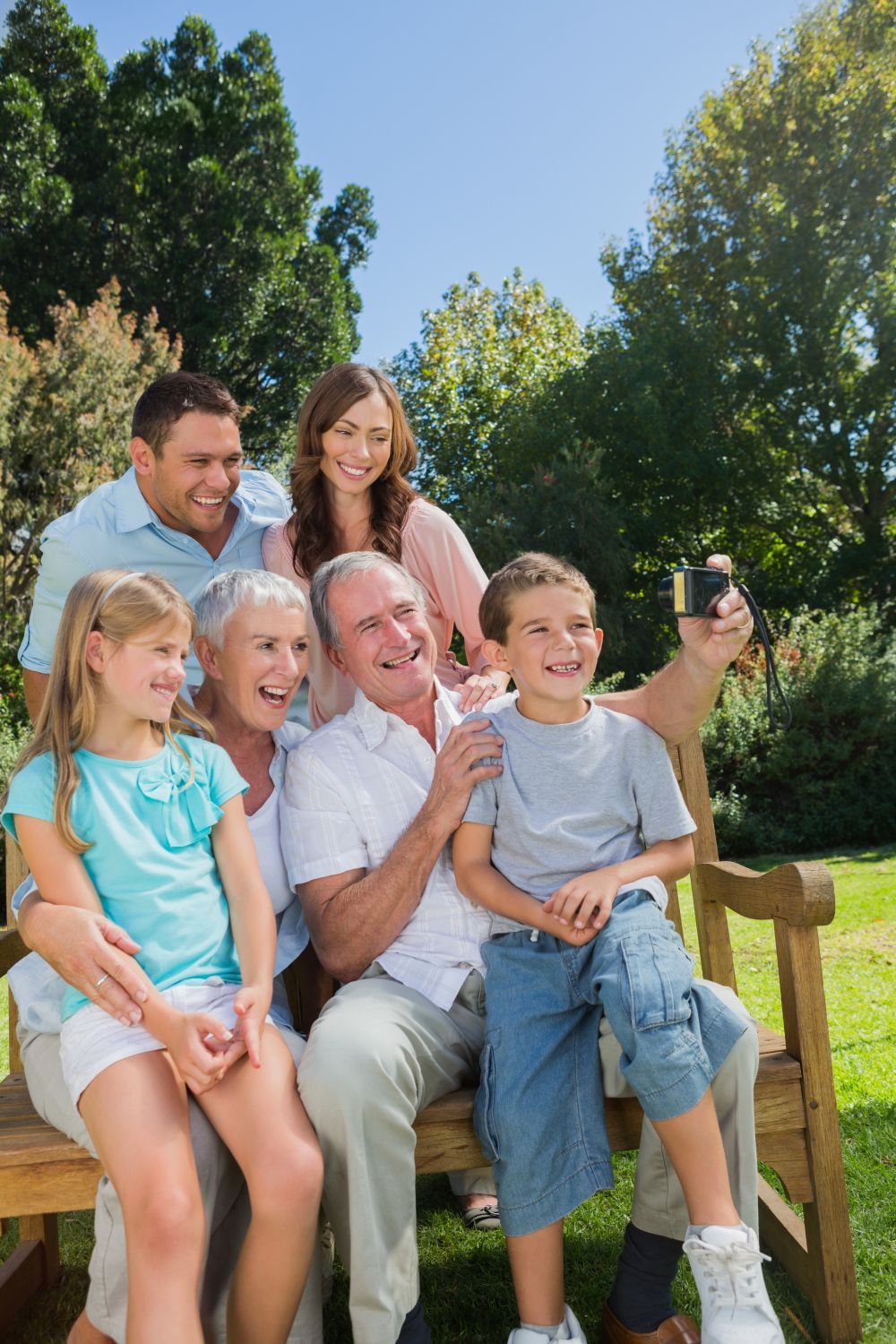 Family poses on a park bench for a selfie. Smiling, sunny day.