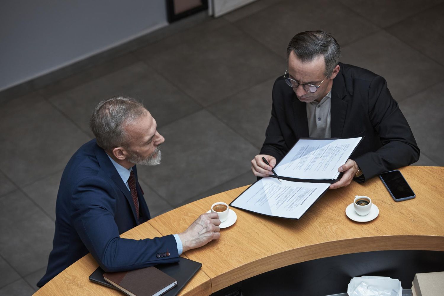 Two men in suits reviewing documents at a table, one drinks coffee.