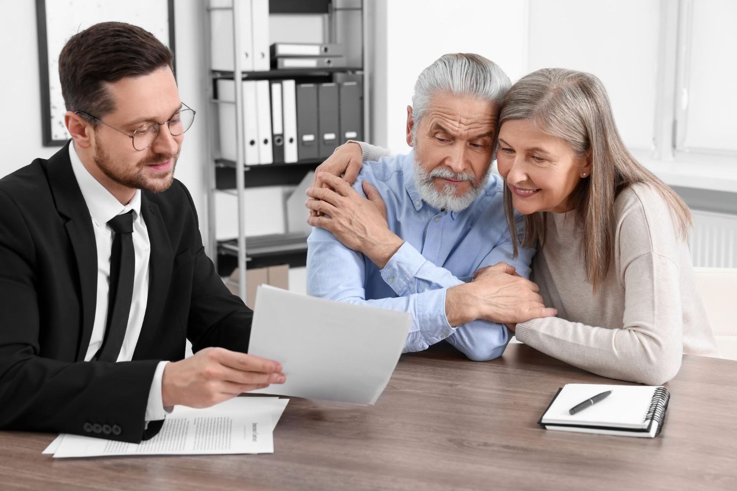 Man in suit explains documents to elderly couple in office; woman comforts man.
