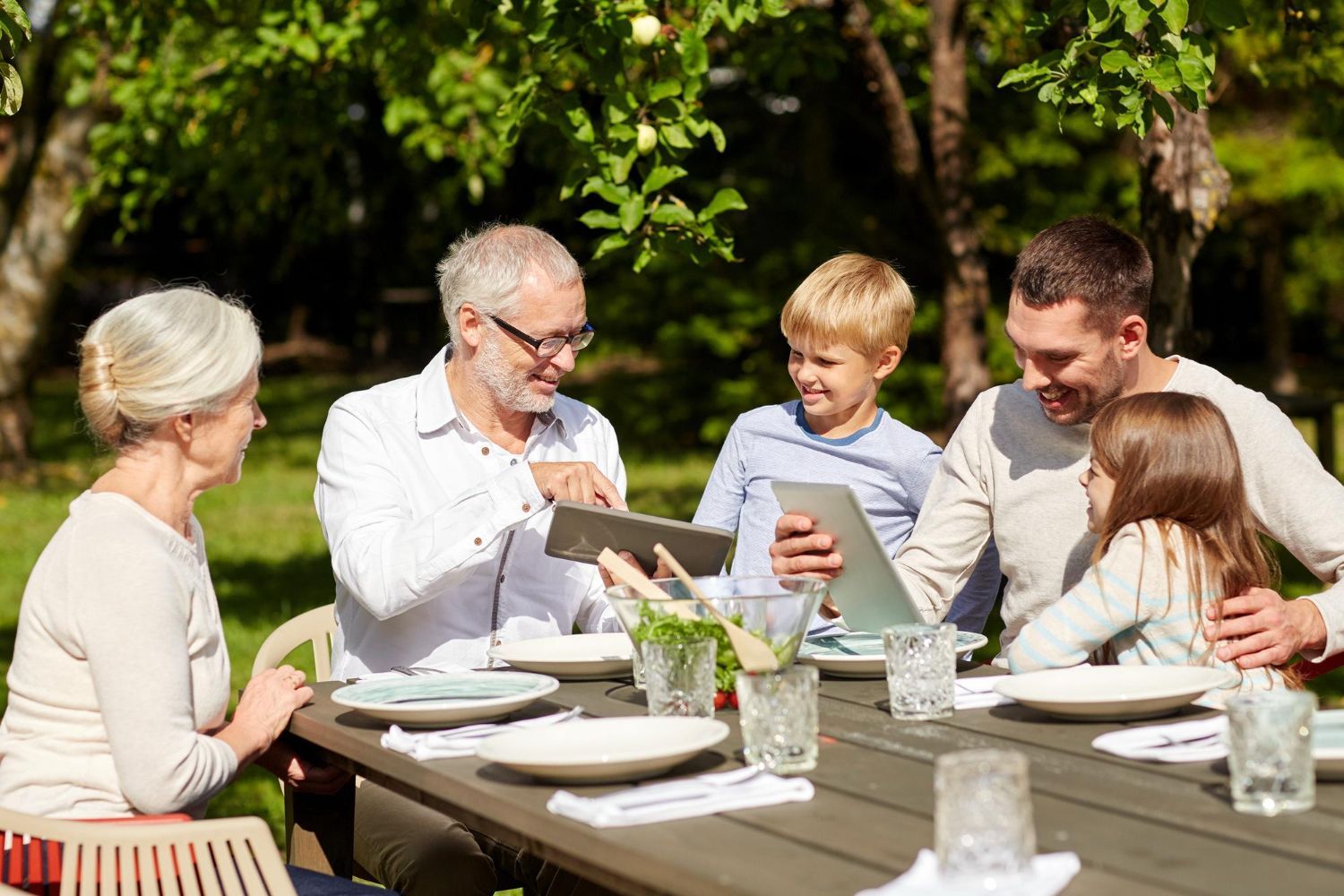 Family, including grandparents, using tablets at an outdoor dining table.