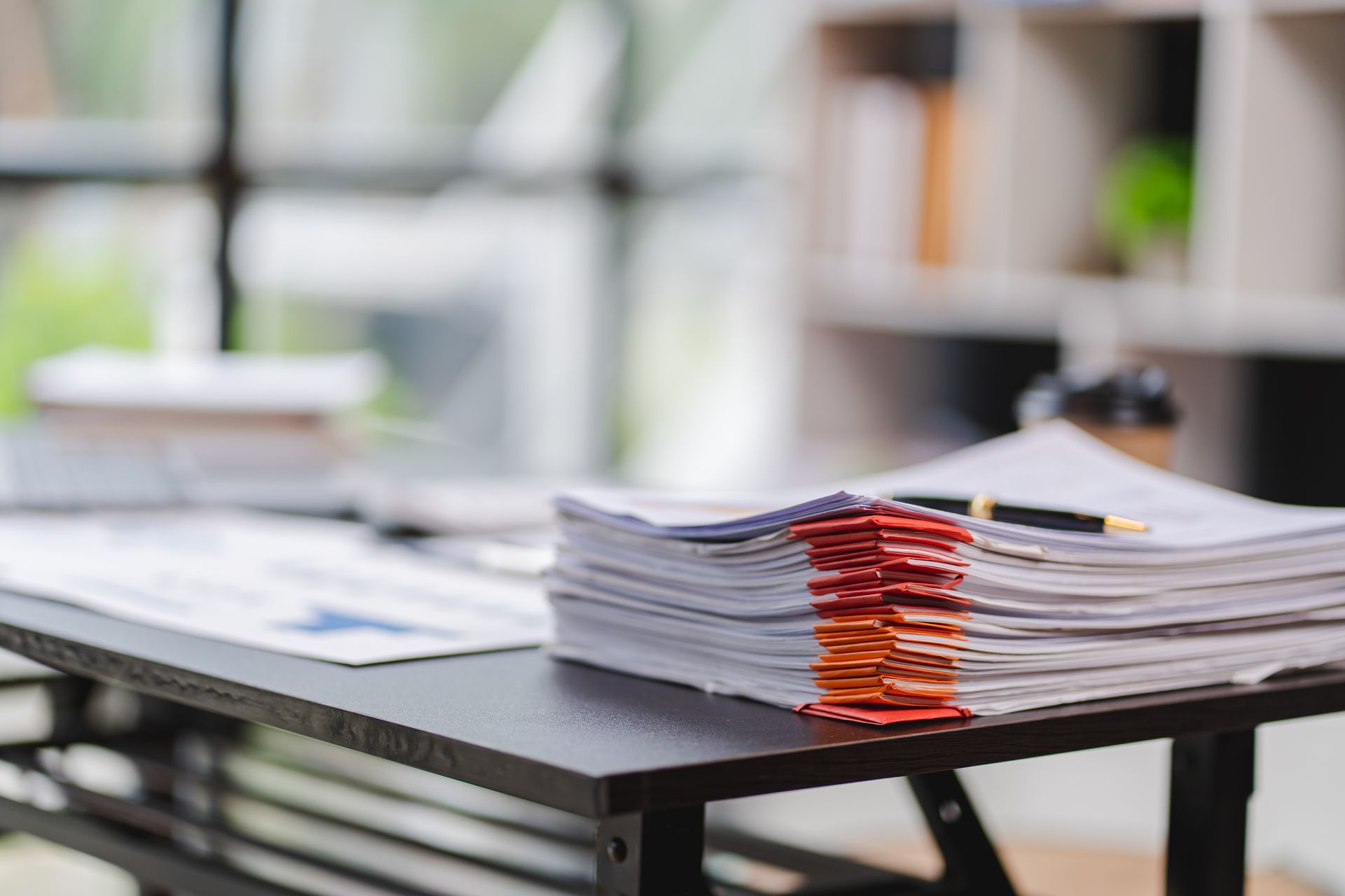 A stack of papers with colored tabs sits on a desk, near a window and a blurred office interior.