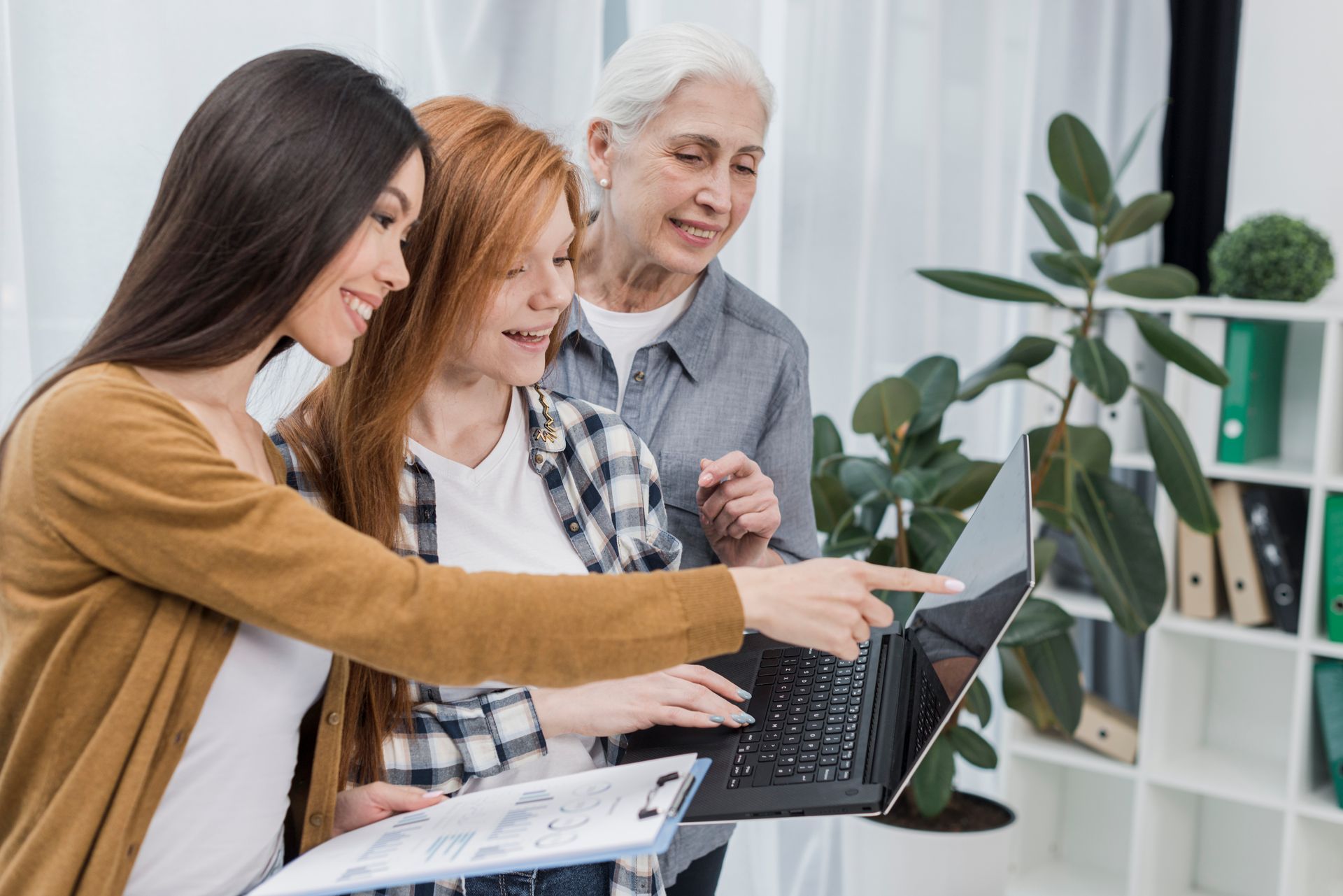 Three women looking at a laptop, one pointing at the screen. Office setting, smiles, various ages.