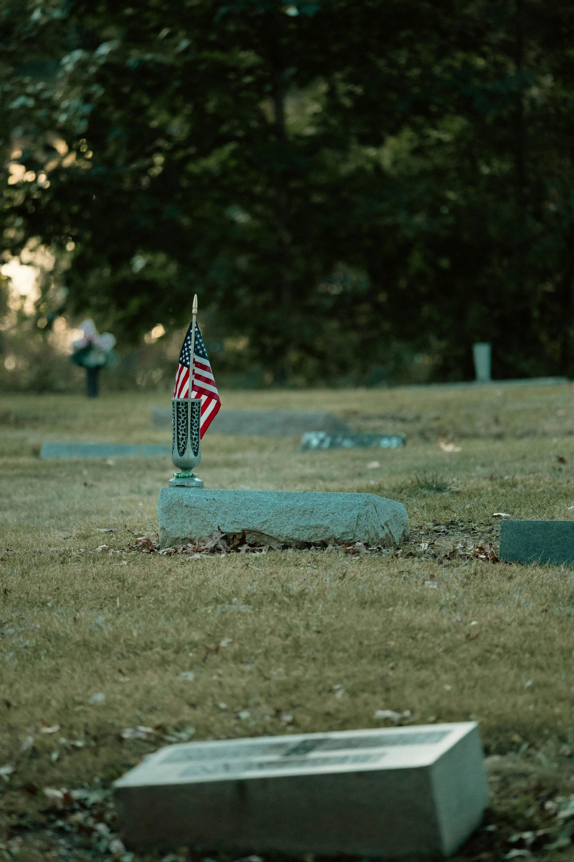 Grave in a cemetery with American flag; dusk lighting; green grass and trees; other headstones.