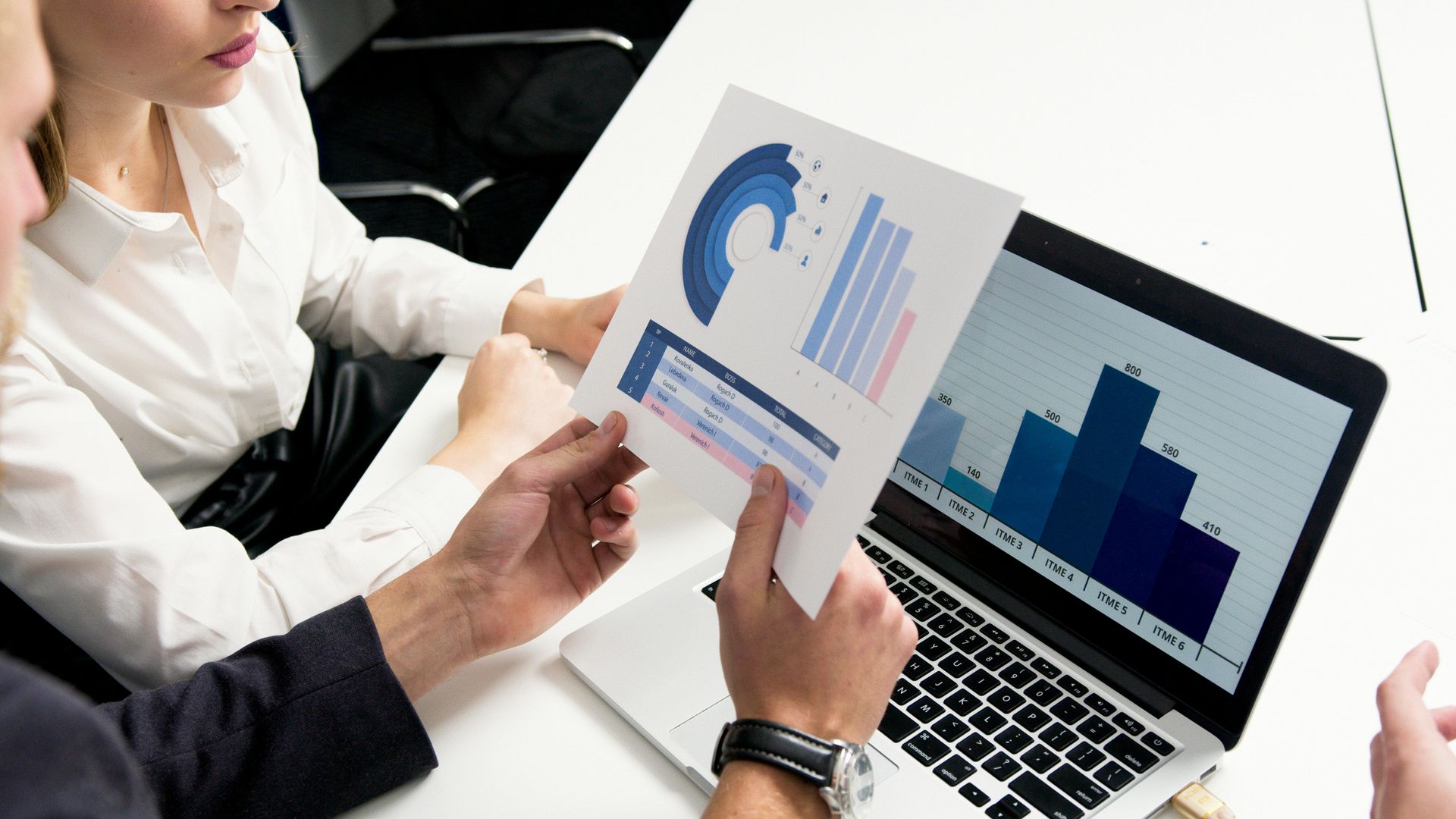 Two people reviewing charts at a laptop on a white desk. One holds a paper chart with blue and pink graphs.