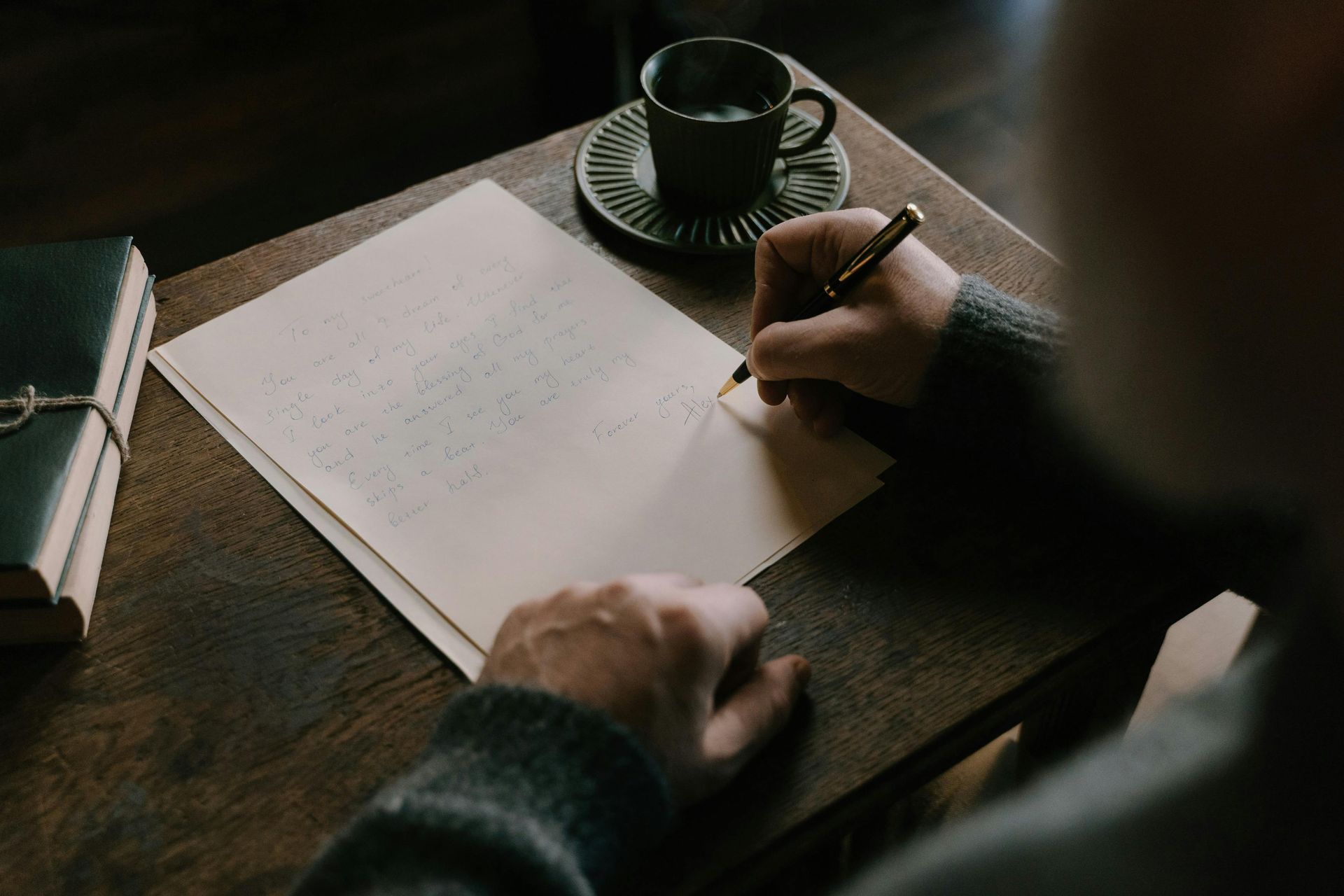 Person writing at a desk with a pen on paper, coffee cup, and a book.