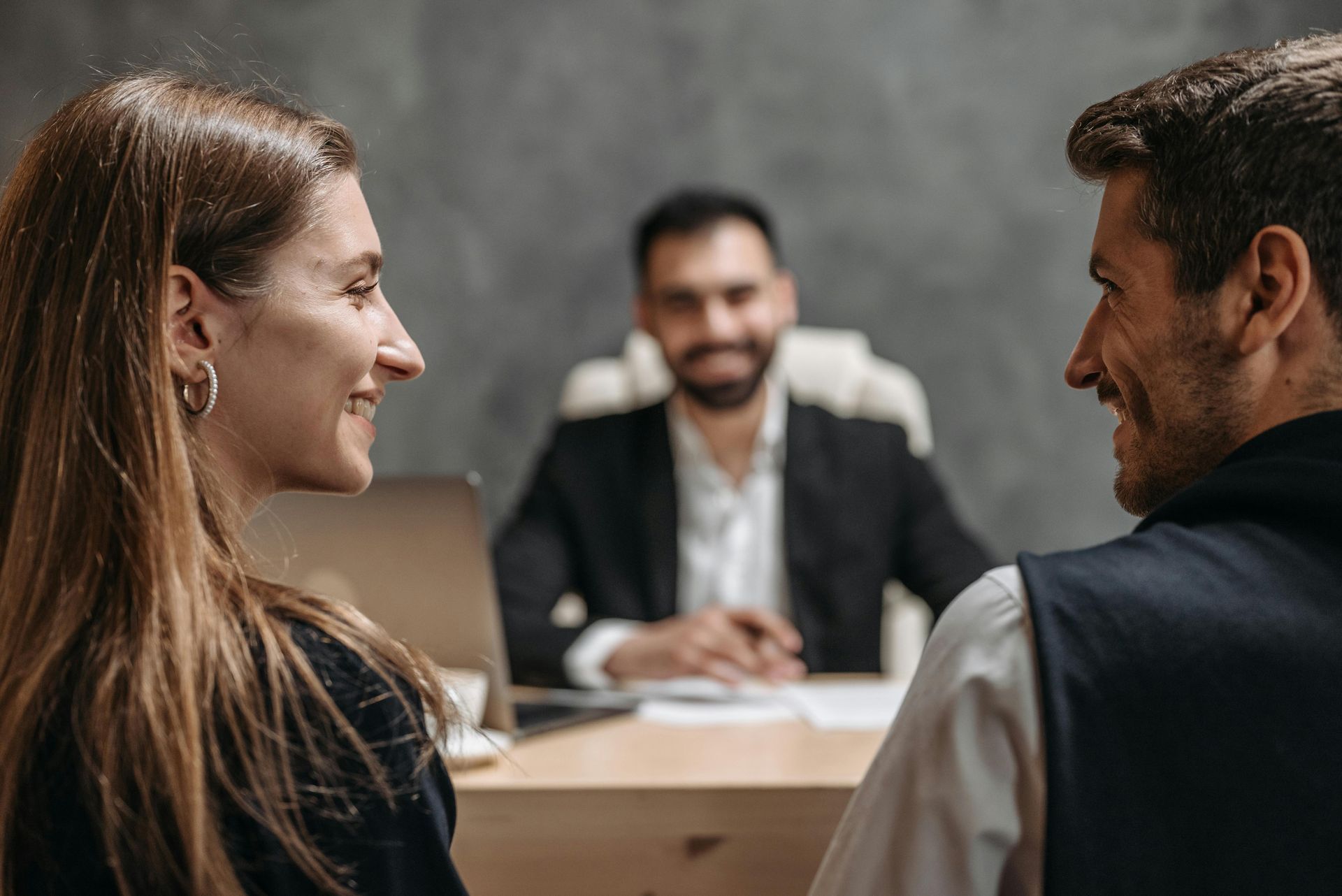Couple smiling at each other across a desk from a smiling man in a suit, indoors.
