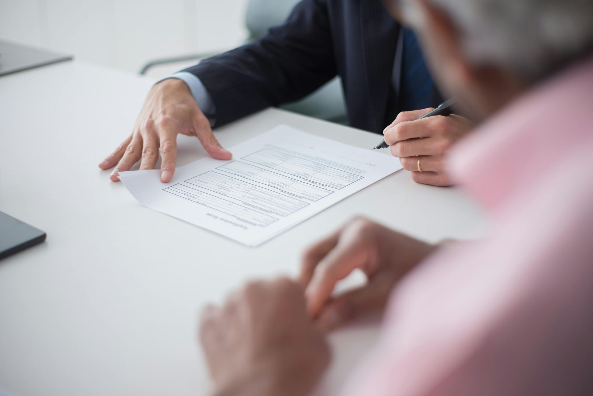 Person signing a document at a white table. A second person looks on, out of focus.