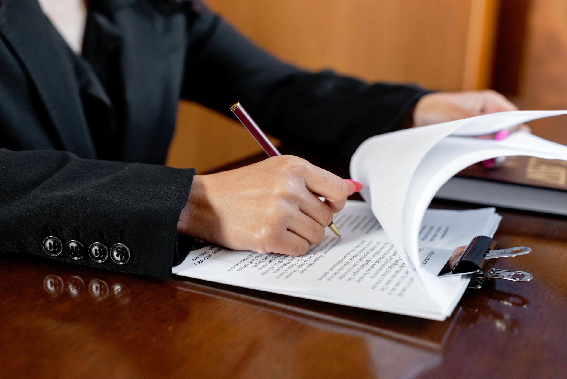 Person in a black suit signing documents with a pen at a wooden desk.