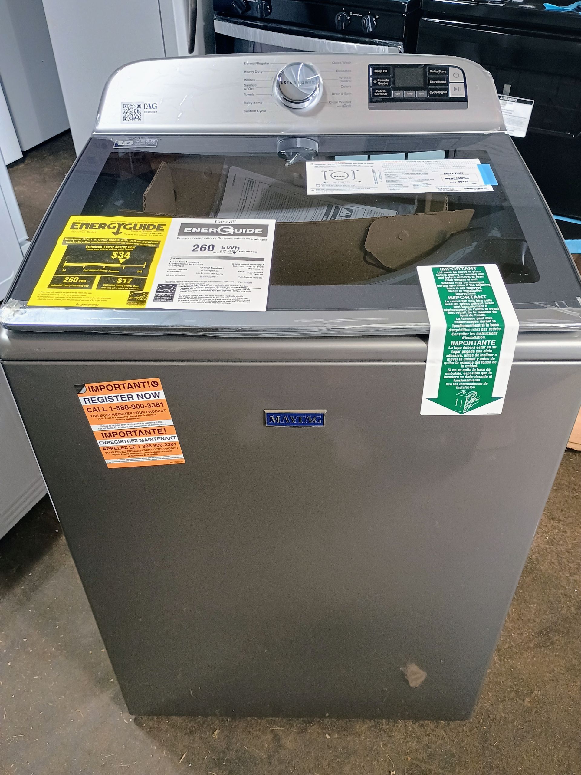 Maytag top-load washing machine with clear lid, several stickers, in a store setting. Gray finish, control panel on top.