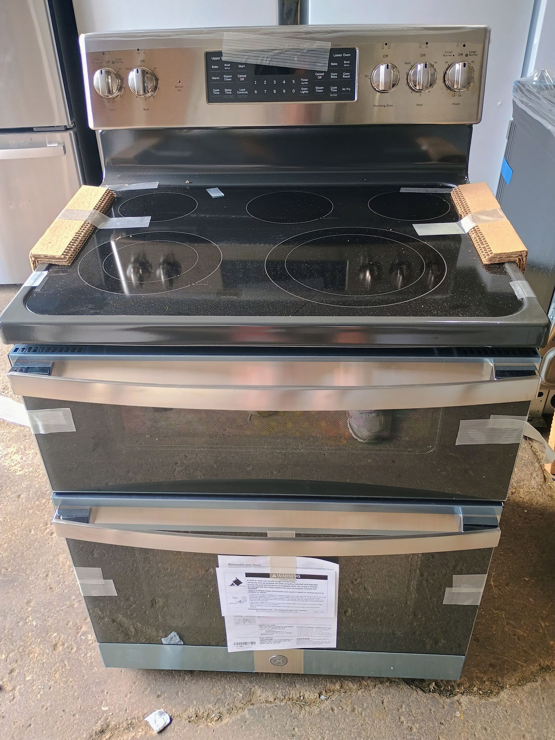 Stainless steel electric range with a black cooktop; protective packaging taped around the oven.