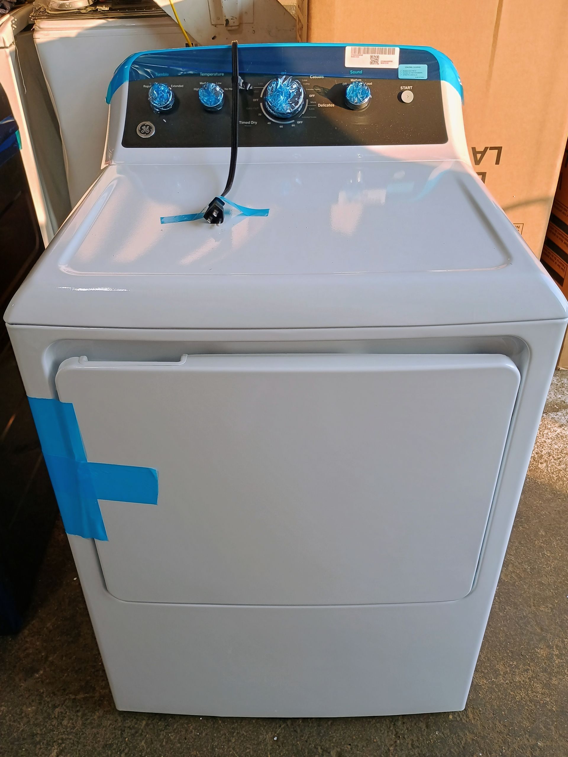 A new, white GE dryer with blue control panel tape. Front view, indoor setting.