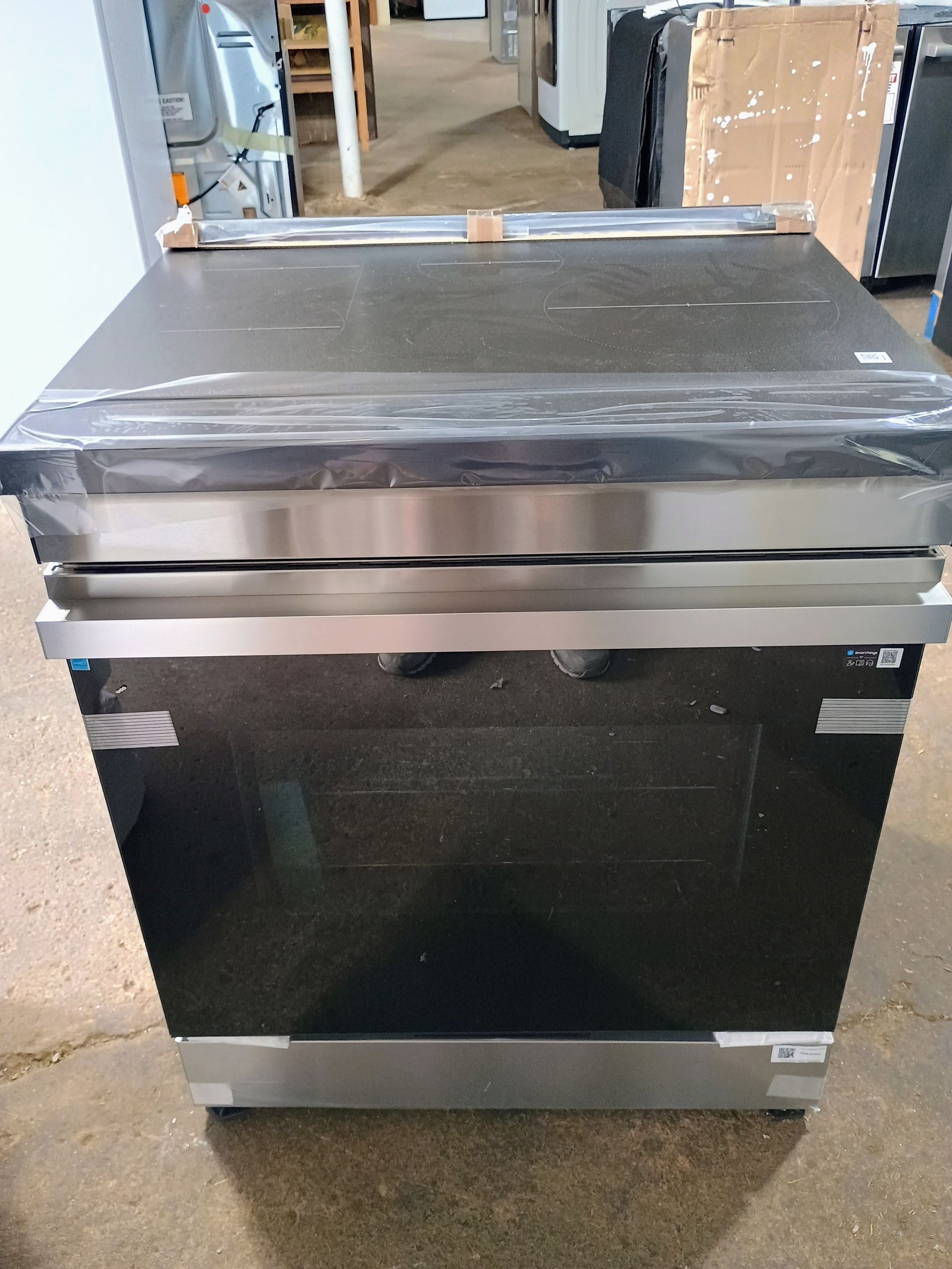 A black and stainless steel oven, wrapped in plastic, stands on a warehouse floor.