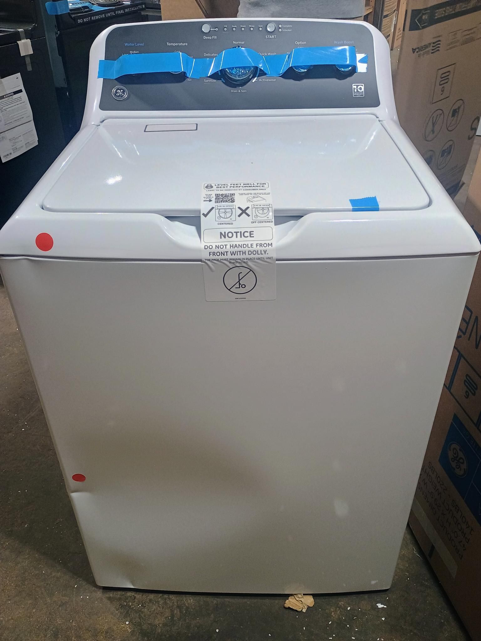 White top-load washing machine with a closed lid. Blue tape and a price tag are attached.