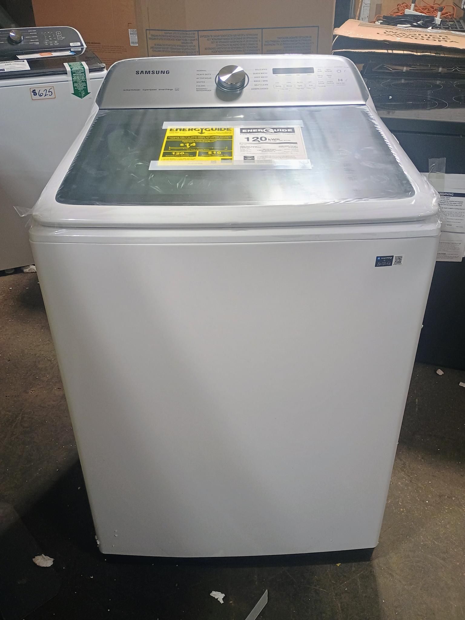 White top-load washing machine with a glass top and silver control panel. A yellow energy sticker is visible on the lid.