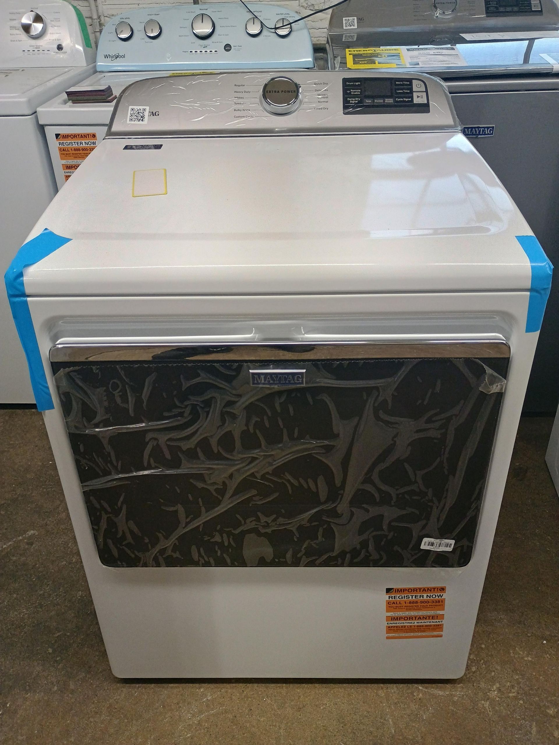 White Samsung dryer with a black door, in a store, still wrapped in plastic.