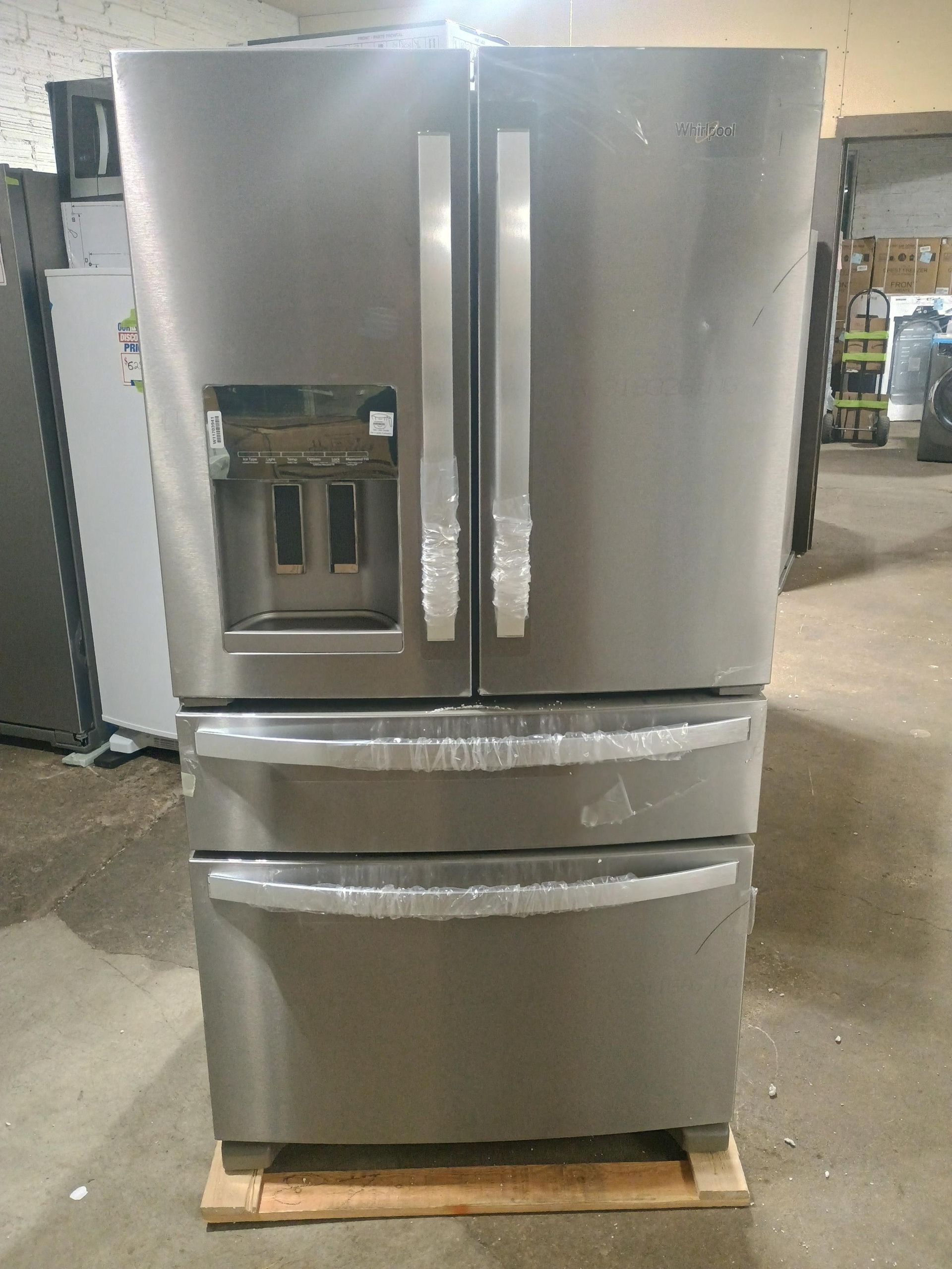 Stainless steel, four-door Whirlpool refrigerator on a pallet, interior shelving visible.