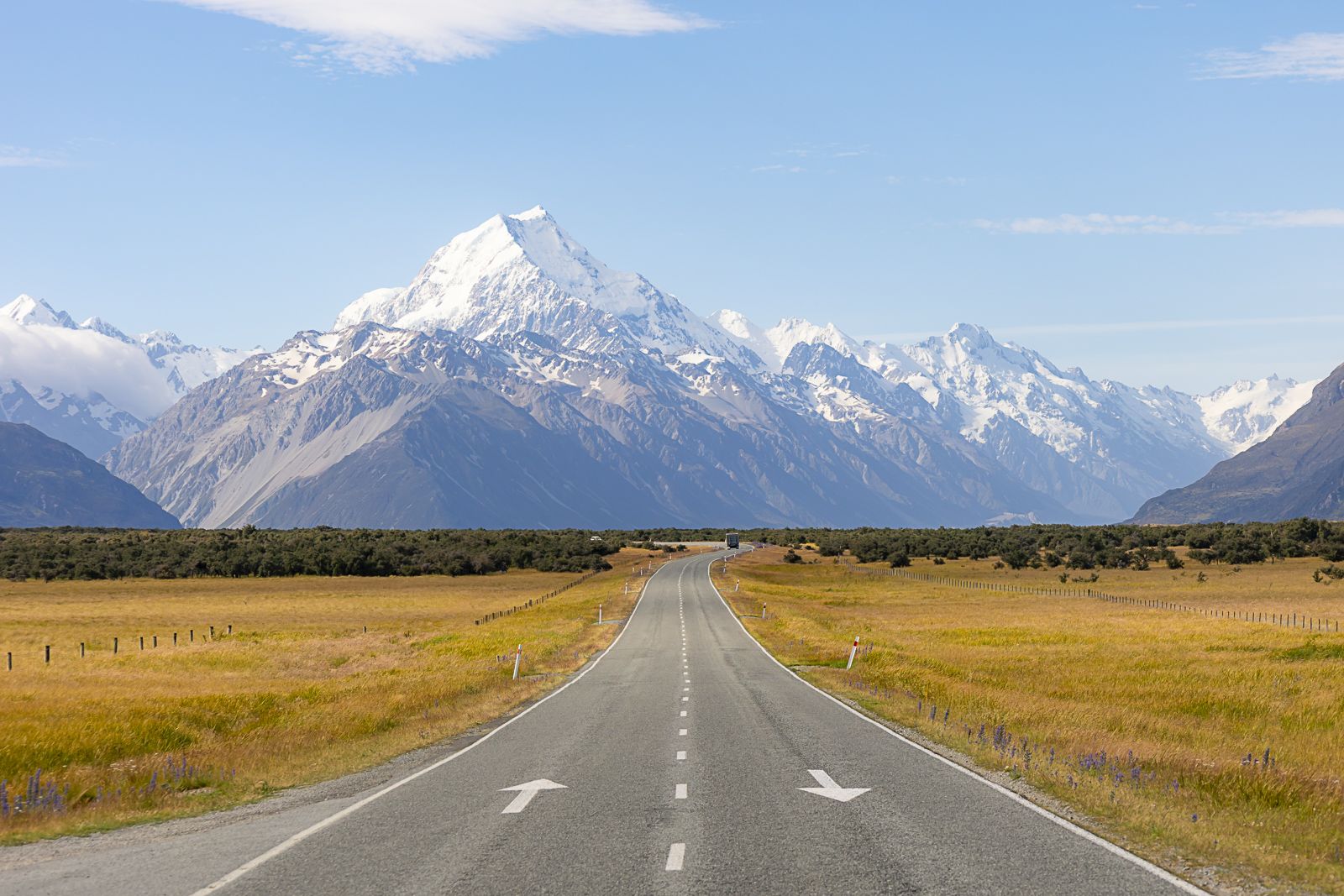 Straight road leading toward snow-capped mountains under a blue sky, with grassy fields on both sides