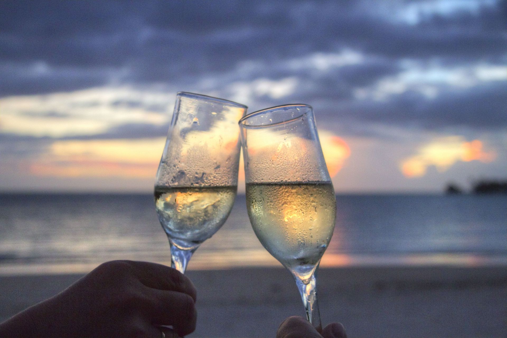Two champagne flutes clink at sunset on a beach, with the ocean and cloudy sky behind.