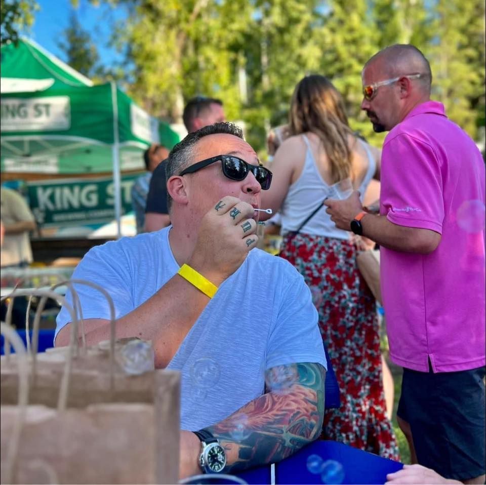 Man in sunglasses smoking, arm tattoos, seated at outdoor event; other people in background.