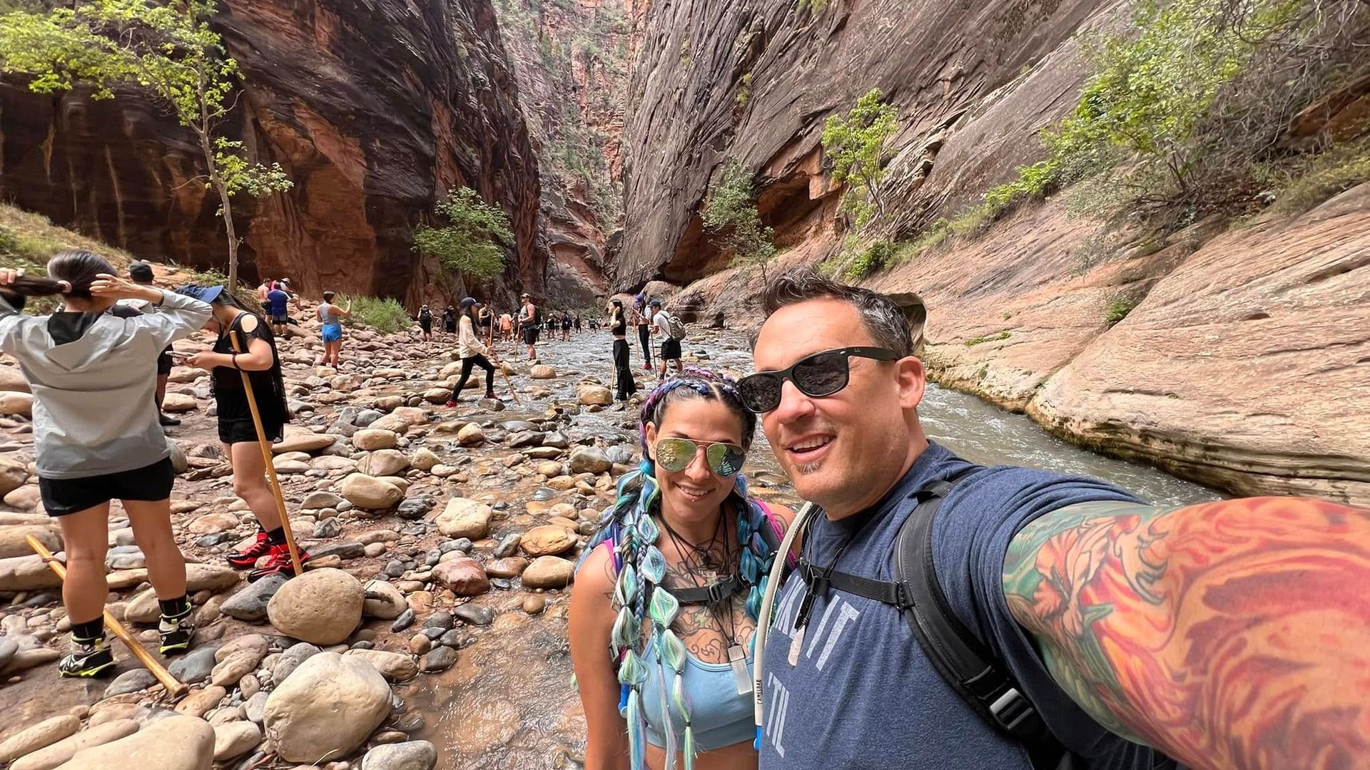 Couple taking a selfie in a narrow canyon with a stream and hikers. Red rock walls, sunny day.