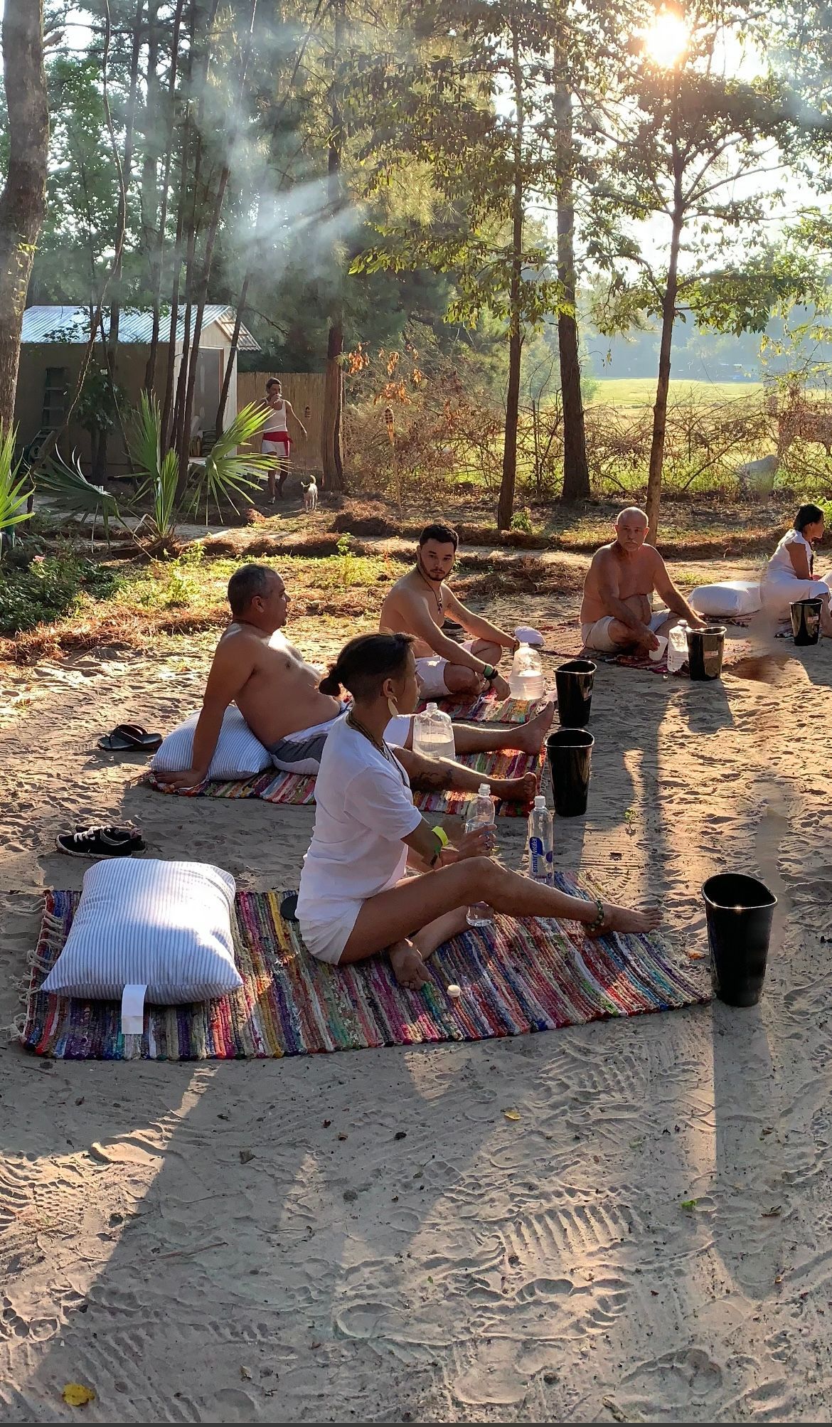 People seated outdoors on mats, engaged in a meditative practice near trees, with sunlight and smoke visible.