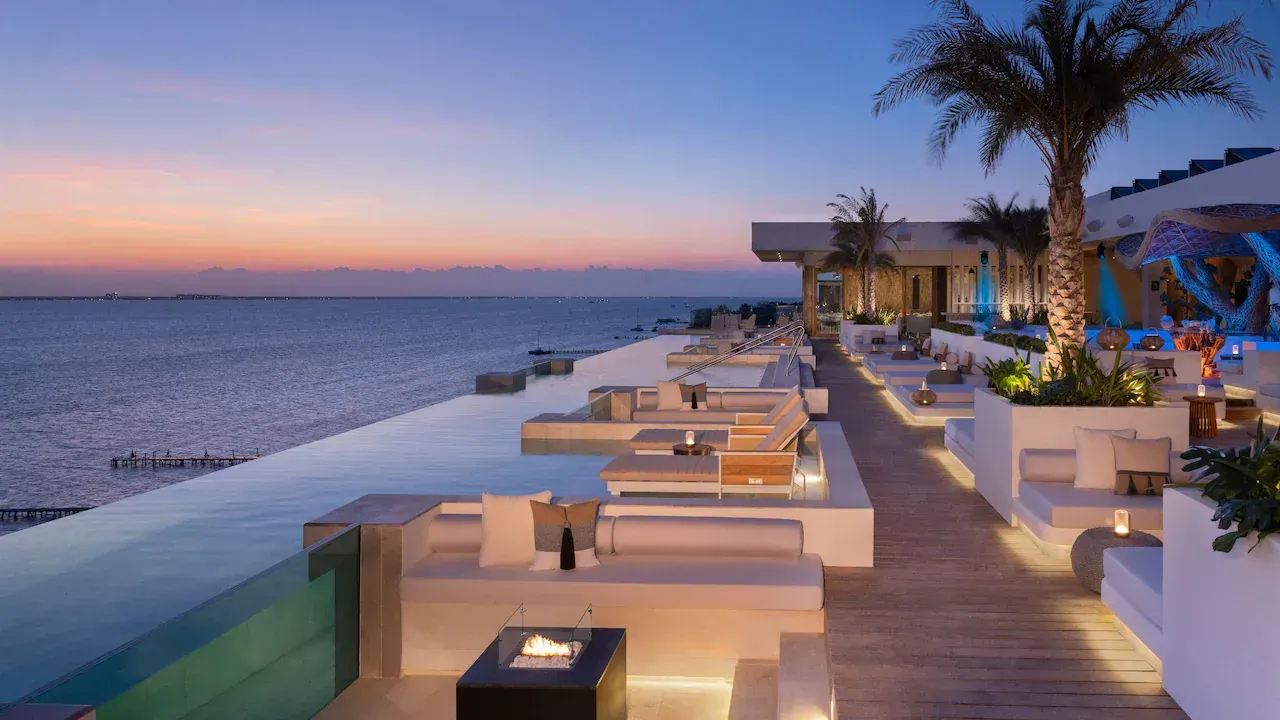 An outdoor lounge area with white seating next to an infinity pool overlooking the ocean at sunset.