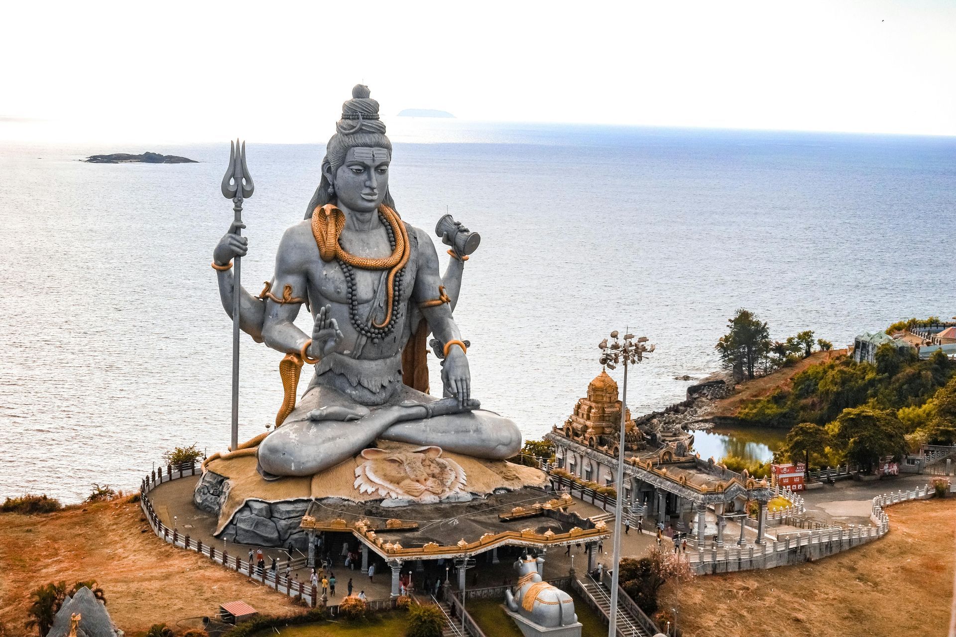 Large statue of Lord Shiva meditating by the ocean. Grey stone figure with trident and peaceful expression.