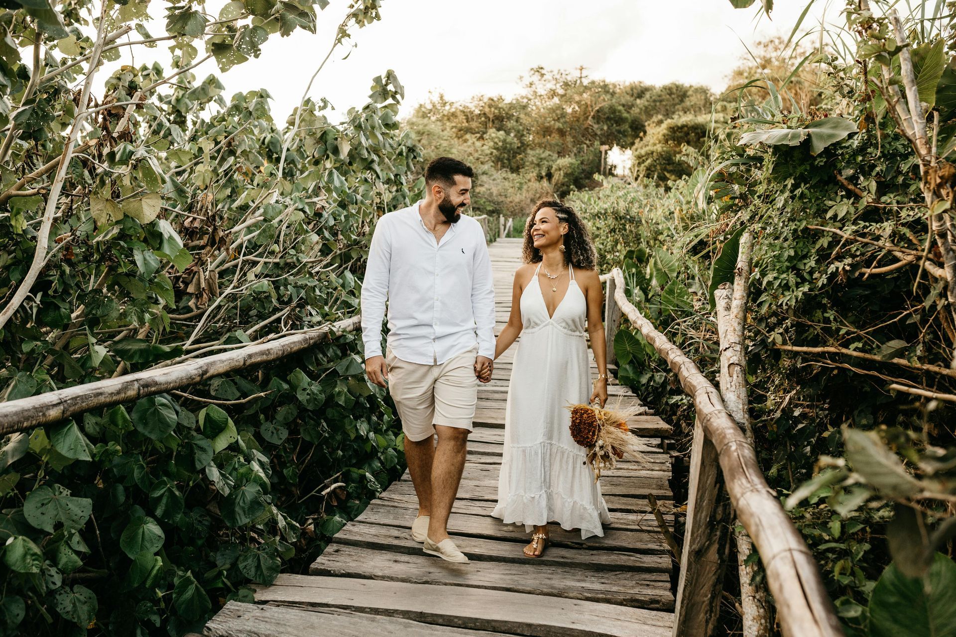 Couple walking on a wooden bridge hand-in-hand, surrounded by greenery, smiling at each other.