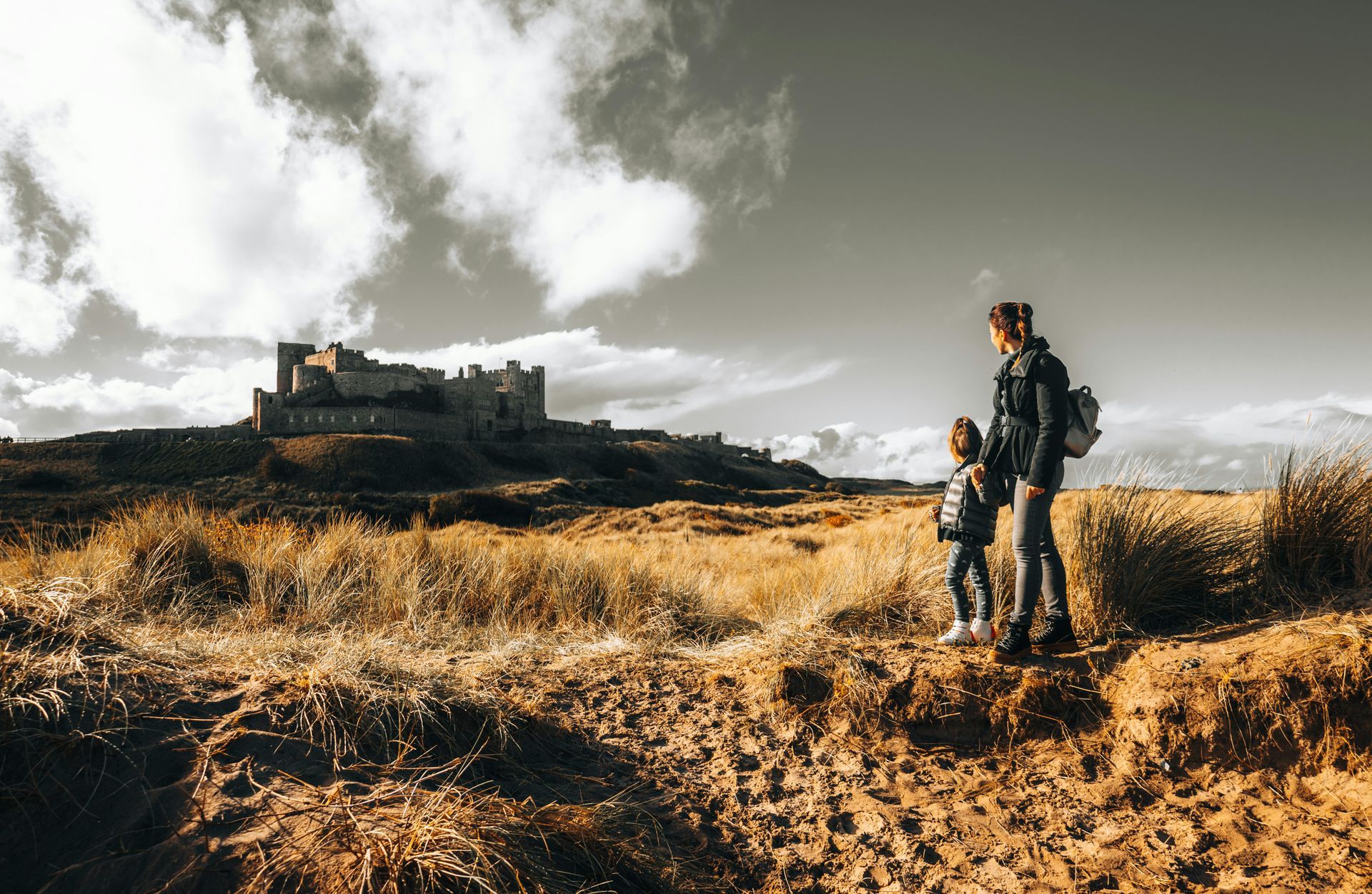 Mother and child stand looking toward a castle on a hill, in a field of brown grass. Cloudy sky.