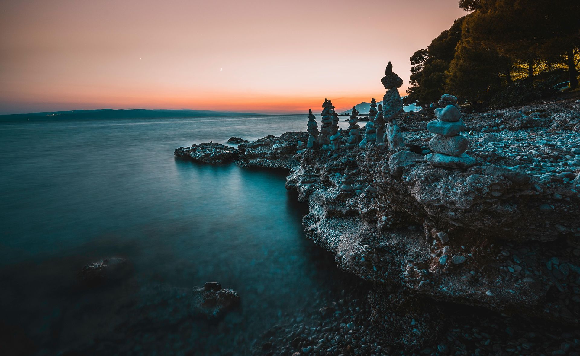 Rocky shoreline at sunset with stacked stone formations. Teal water, orange sky.