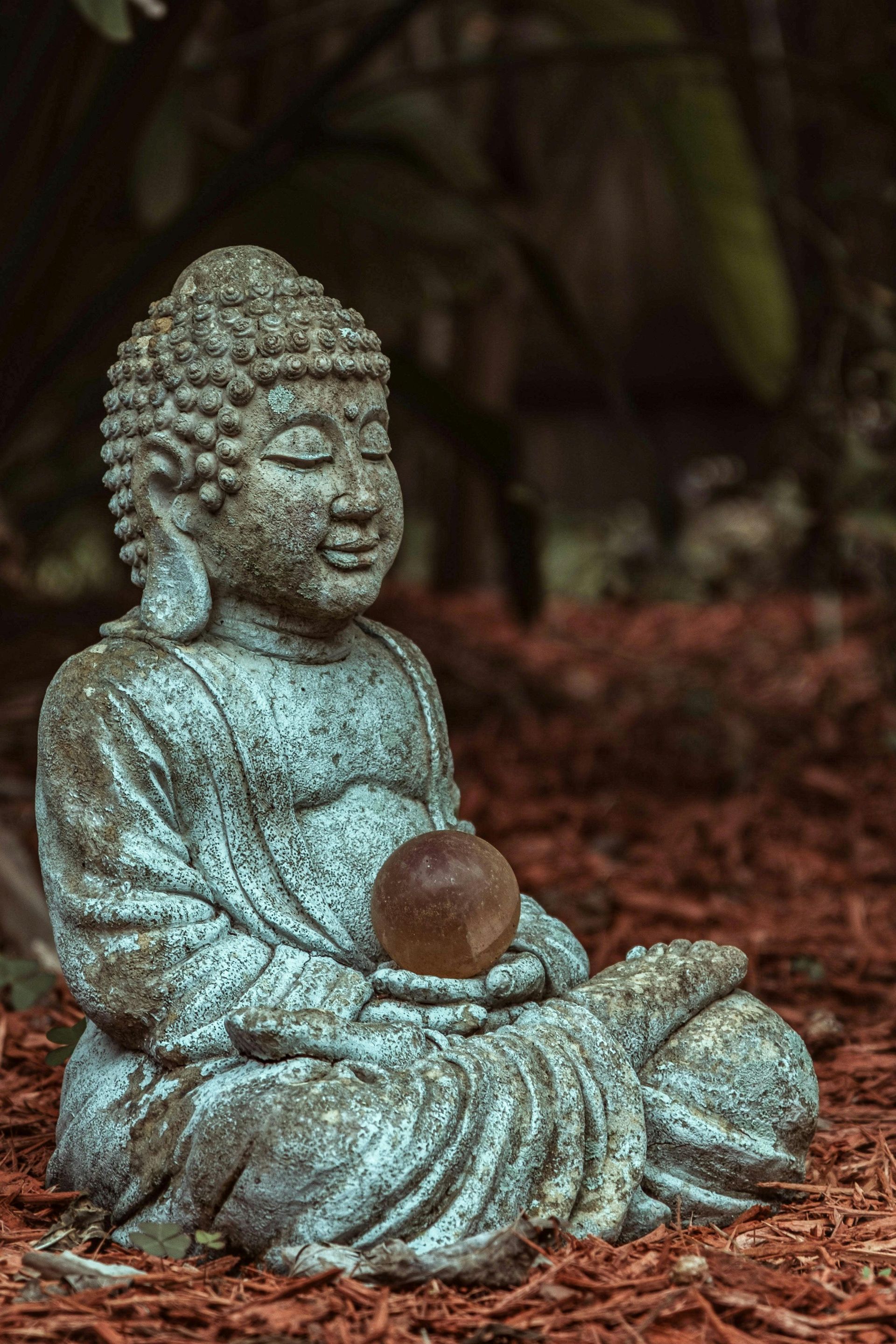 Stone Buddha statue seated in meditation, holding a brown orb, set in a garden.