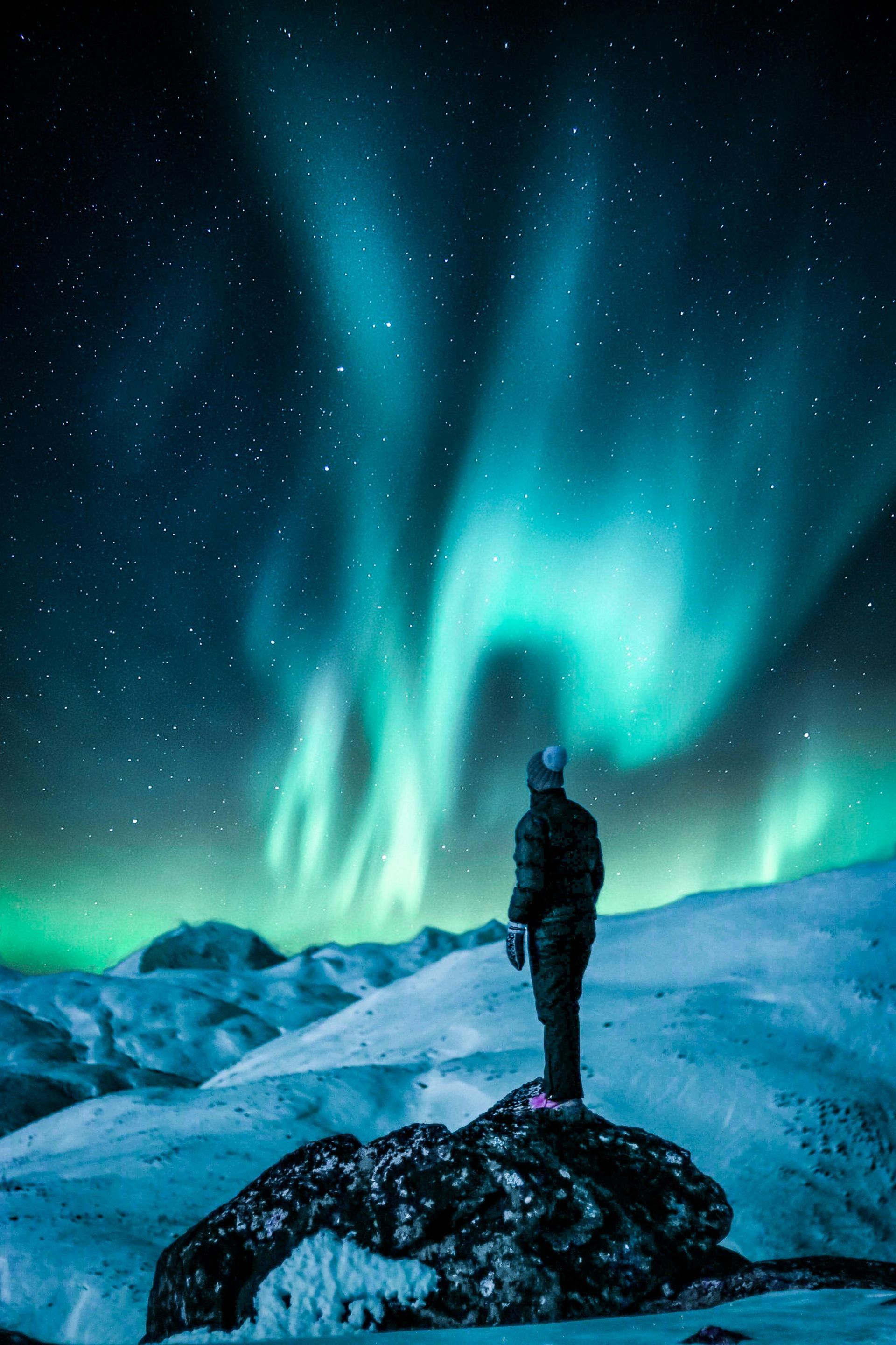 Person stands on snowy rock, gazing at vibrant green and blue aurora borealis in the night sky.