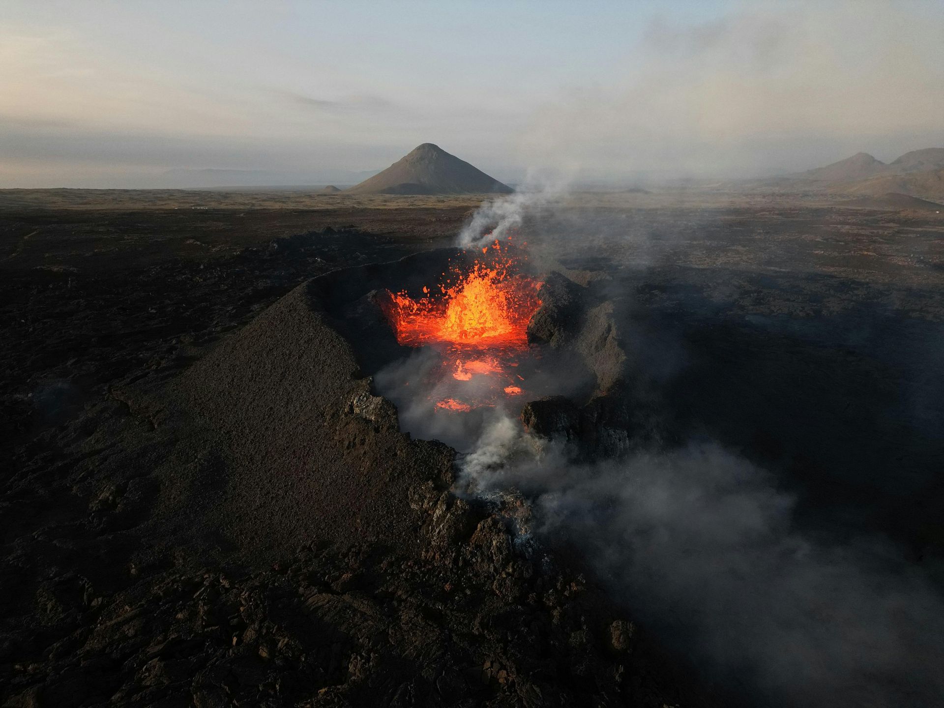 Volcano erupting, spewing lava and smoke into the air; brown landscape, gray sky.