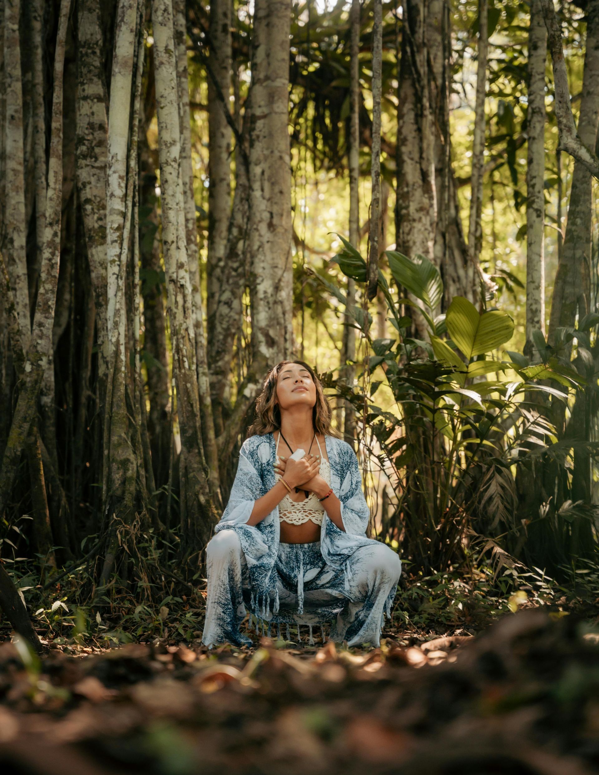 Woman in tie-dye outfit meditates in forest, gazing upwards, hands at chest. Sunlight filters through trees.