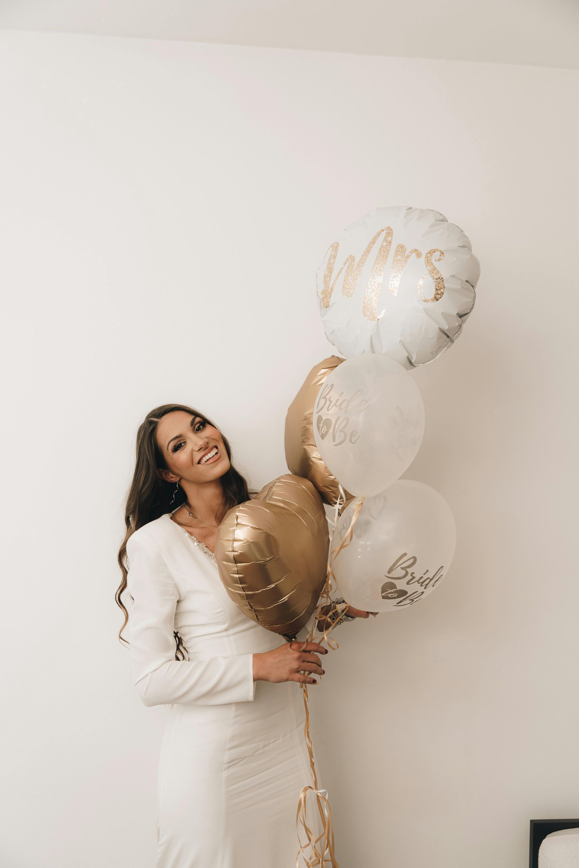 Woman in white dress smiles, holding gold and white balloons with