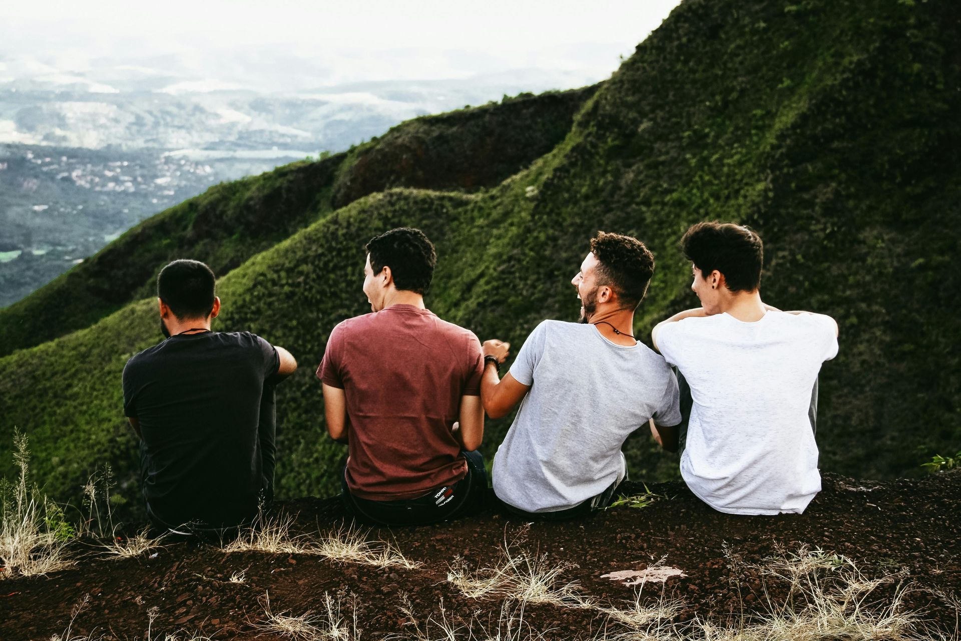 Four young men sit on a cliff, overlooking a green mountain range.