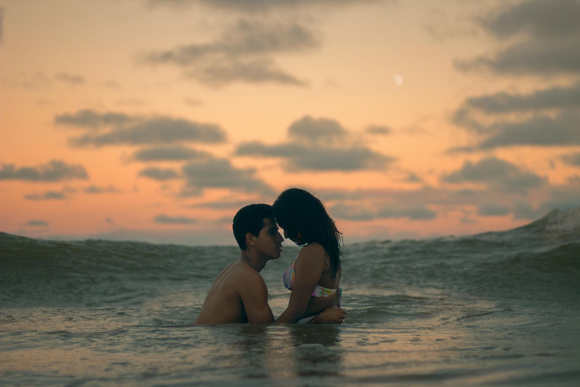 Couple in ocean at sunset, faces close. Orange sky, calm water.