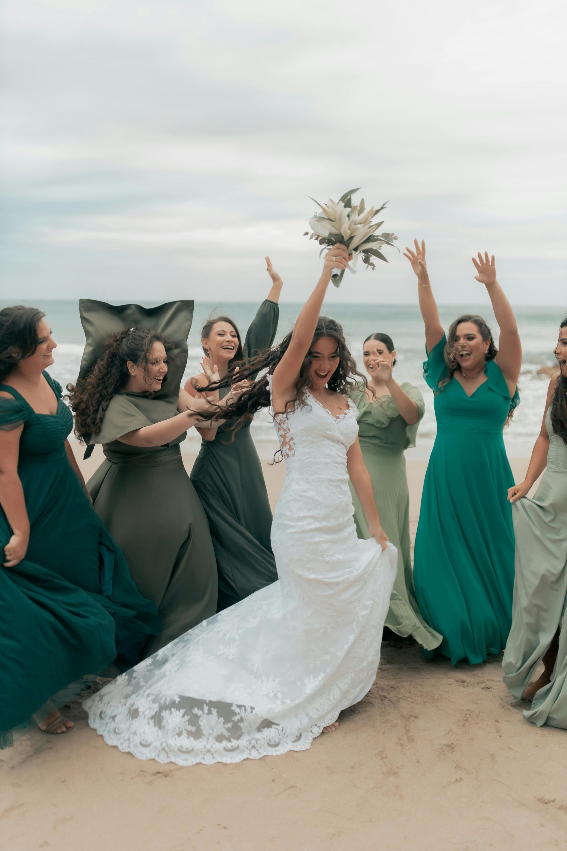 Bride and bridesmaids celebrate on beach, bride in white dress, others in green dresses, all cheering.